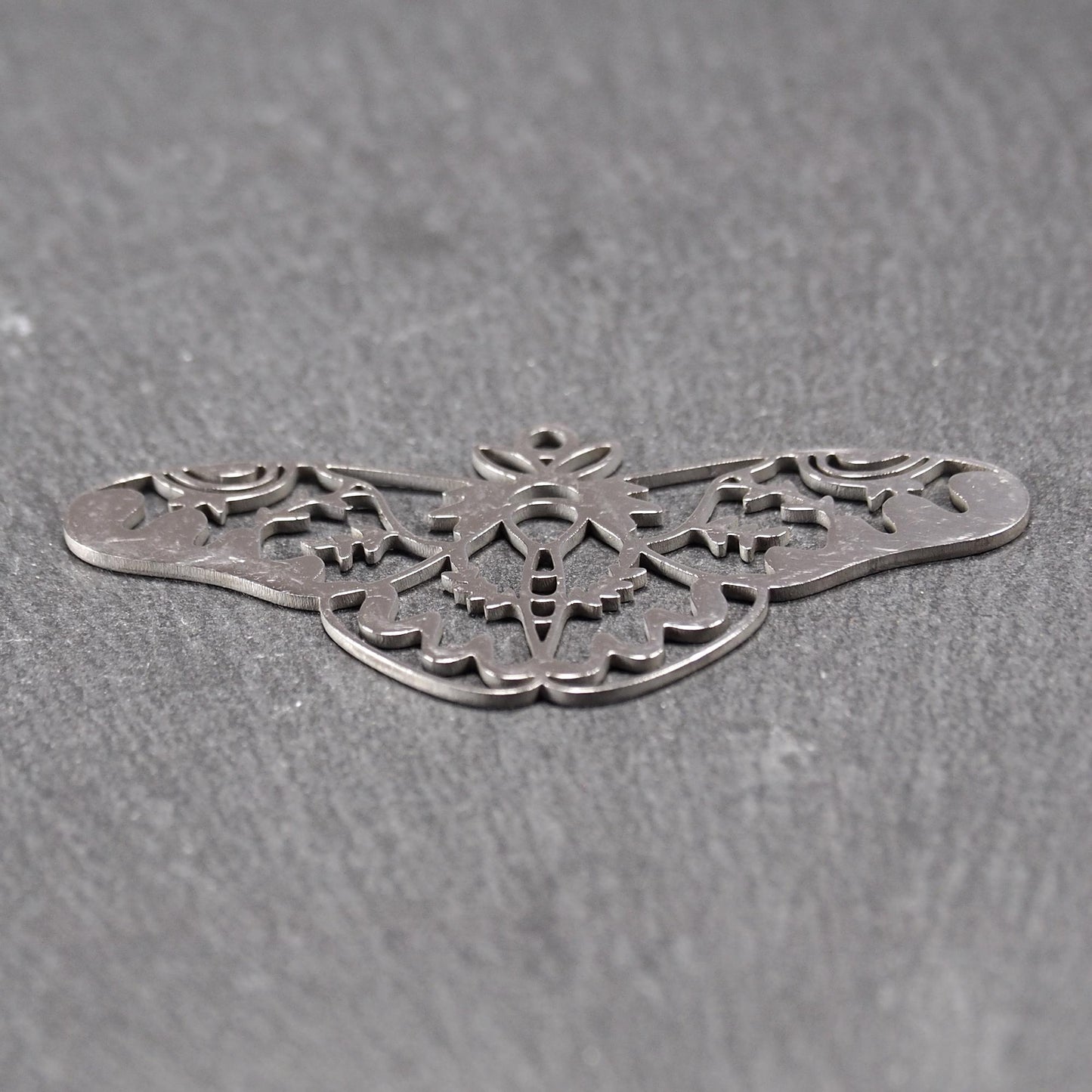Anhänger / Verbinder in Mottenform aus poliertem silberfarbenem Edelstahl, filigranes durchbrochenes Ornament-Flügeldesign mit integrierter Öse oben.