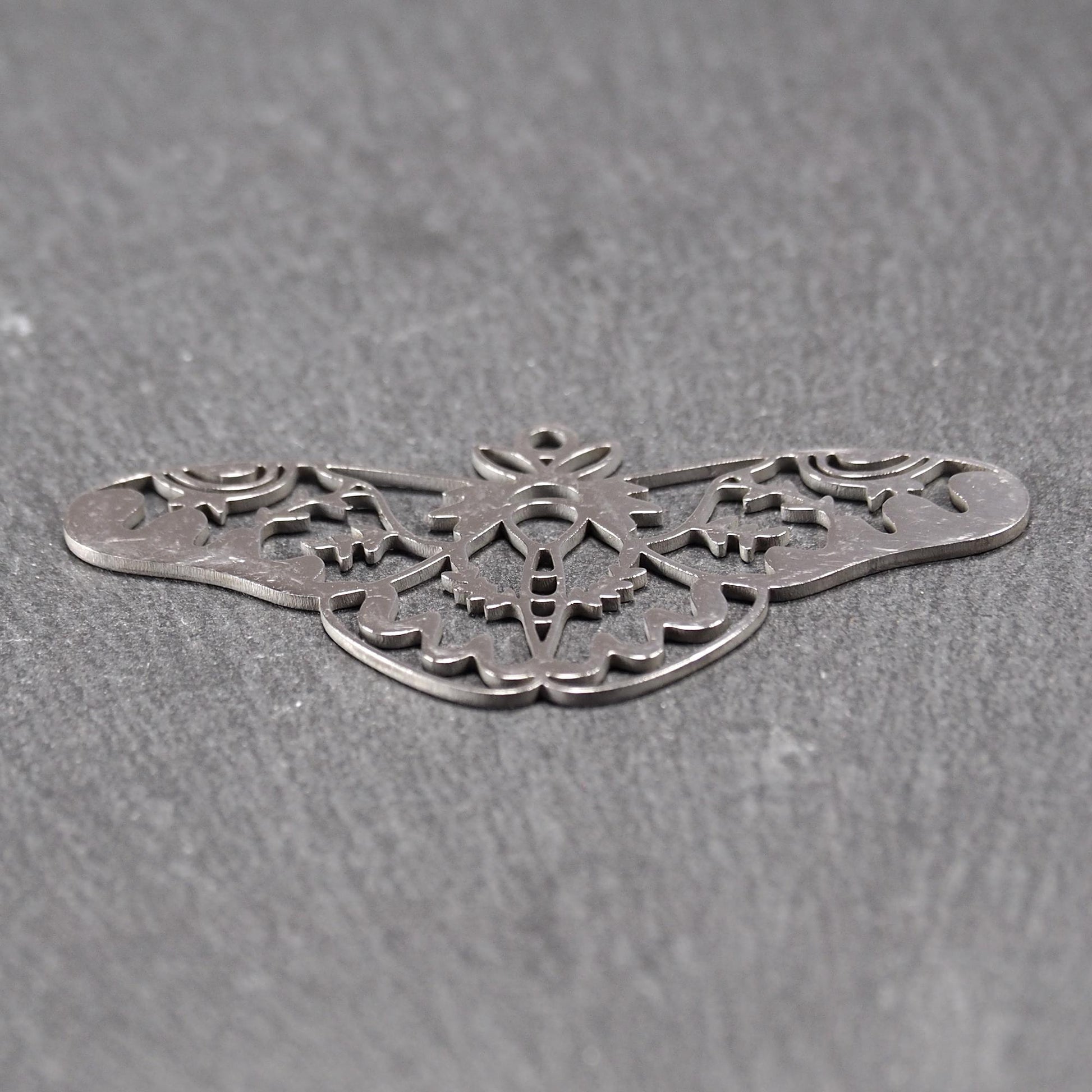Anhänger / Verbinder in Mottenform aus poliertem silberfarbenem Edelstahl, filigranes durchbrochenes Ornament-Flügeldesign mit integrierter Öse oben.