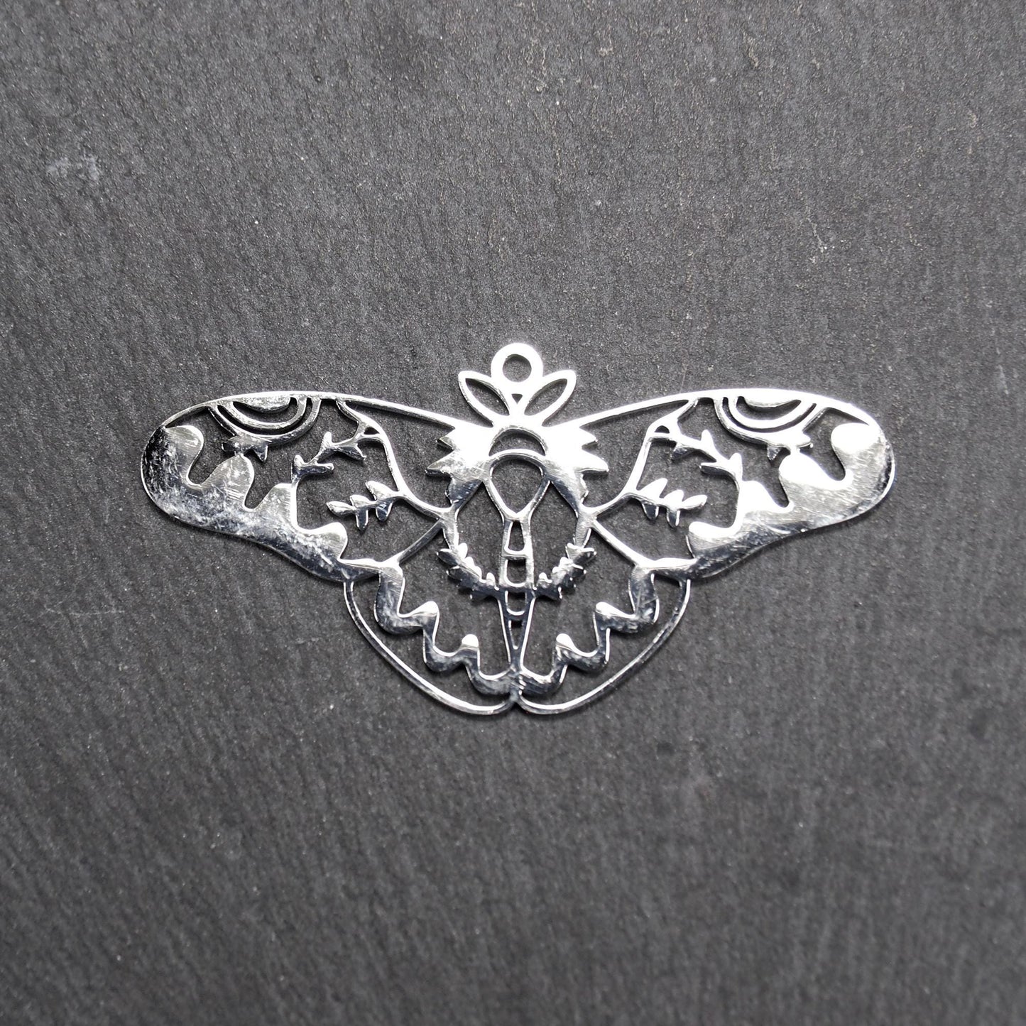 Anhänger / Verbinder in Mottenform aus poliertem silberfarbenem Edelstahl, filigranes durchbrochenes Ornament-Flügeldesign mit integrierter Öse oben.