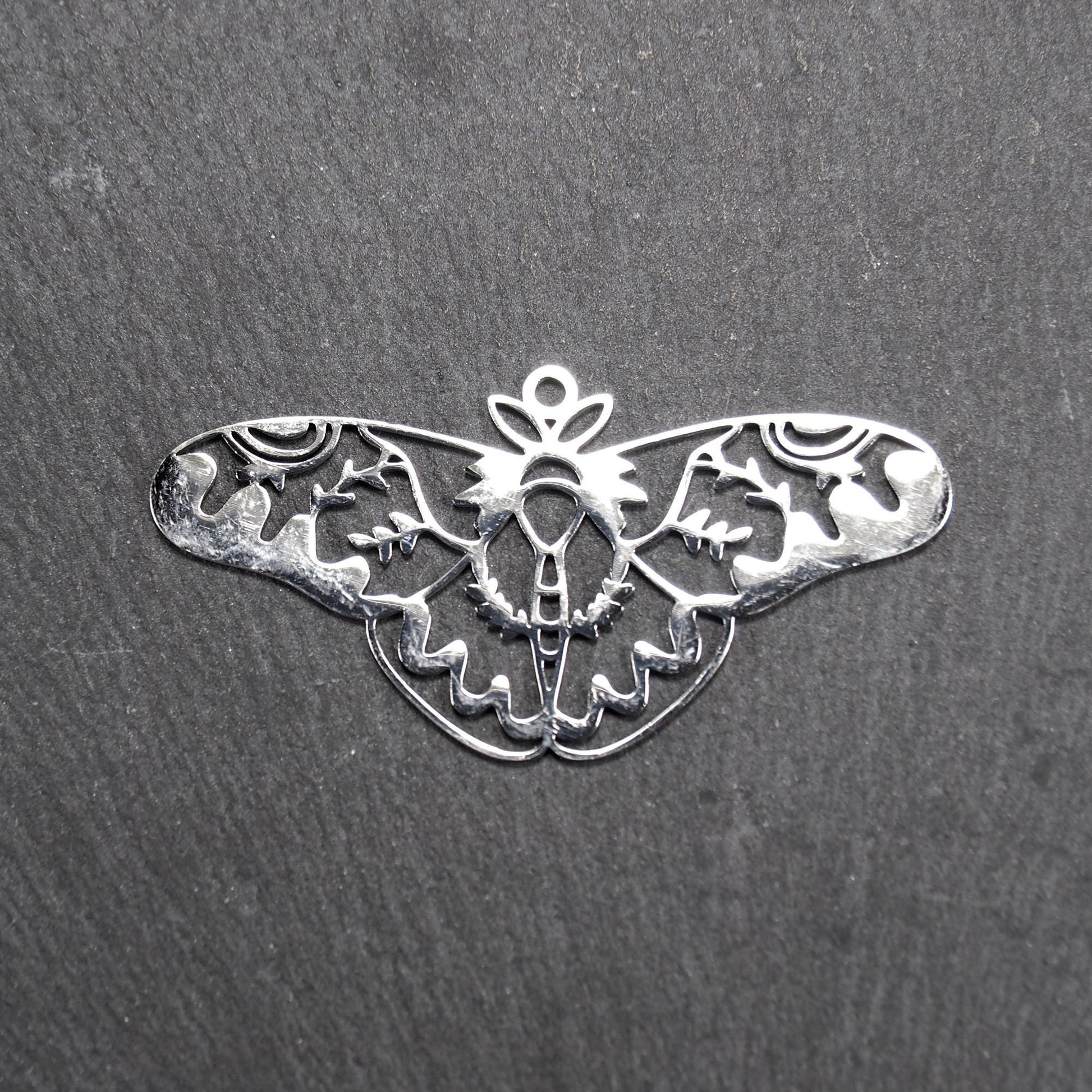 Anhänger / Verbinder in Mottenform aus poliertem silberfarbenem Edelstahl, filigranes durchbrochenes Ornament-Flügeldesign mit integrierter Öse oben.