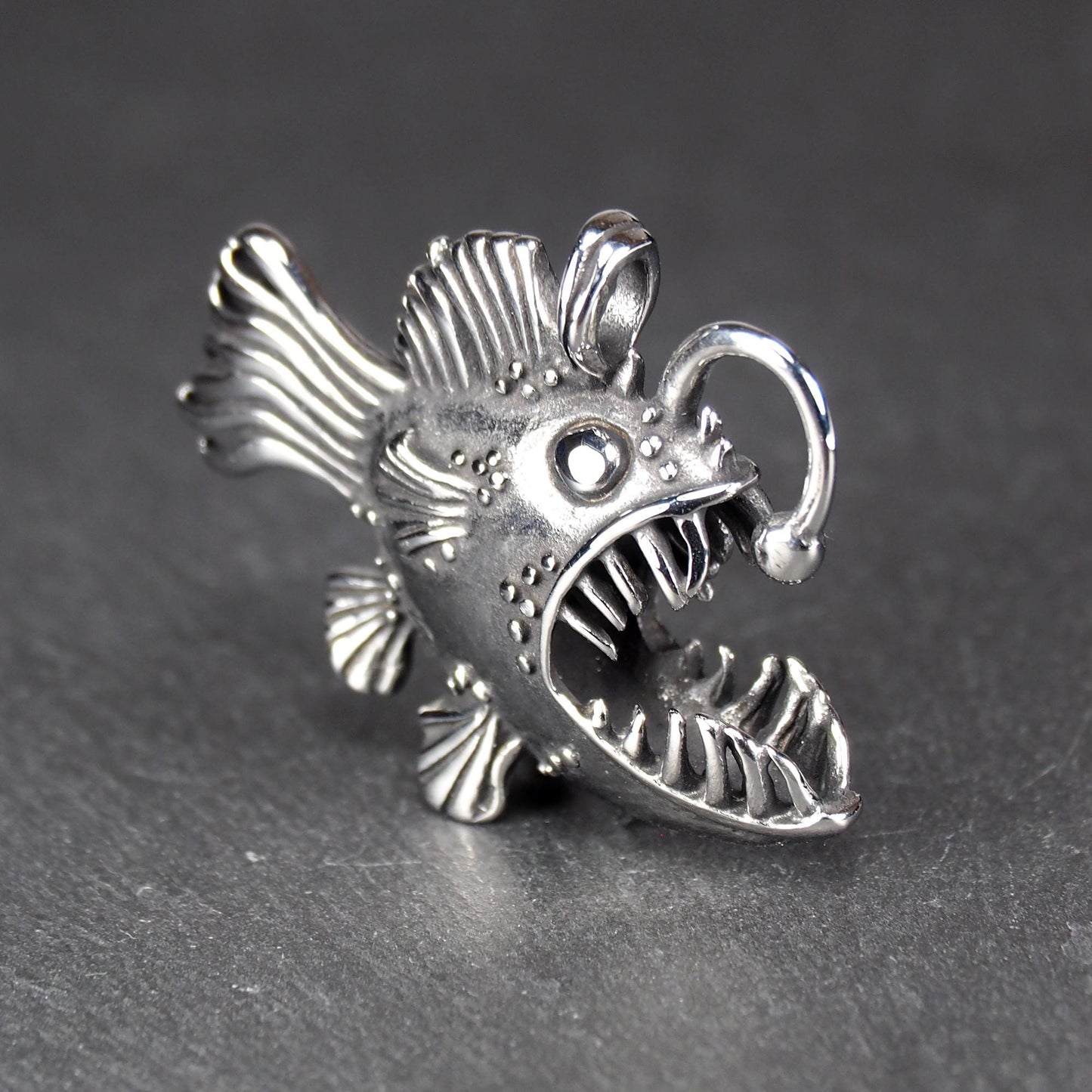 3D-Anhänger Anglerfisch, silberglänzend, mit geöffnetem Maul und dolchartigen Zähnen, geriffelten Flossen, punktiertem Körper und Öse.