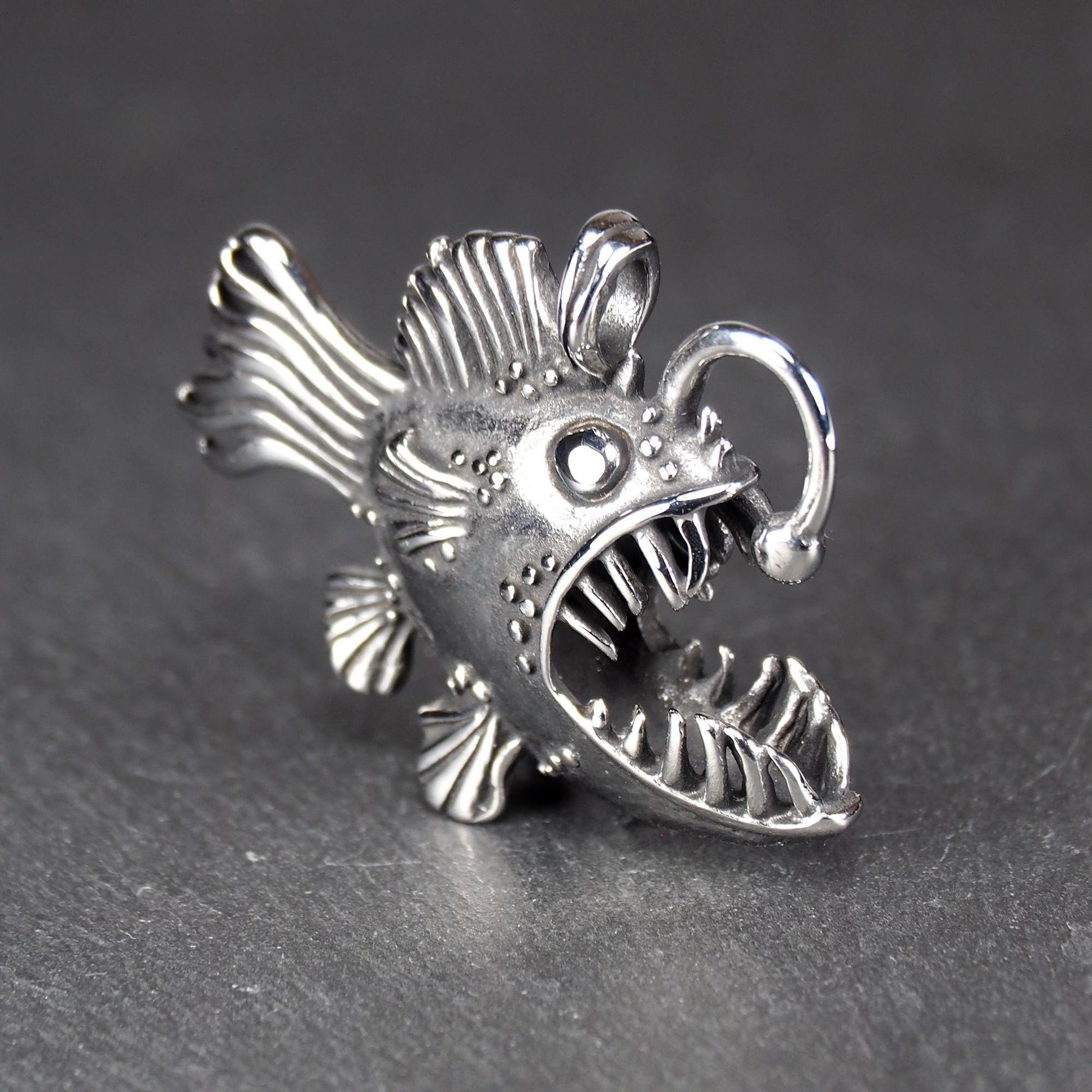 3D-Anhänger Anglerfisch, silberglänzend, mit geöffnetem Maul und dolchartigen Zähnen, geriffelten Flossen, punktiertem Körper und Öse.