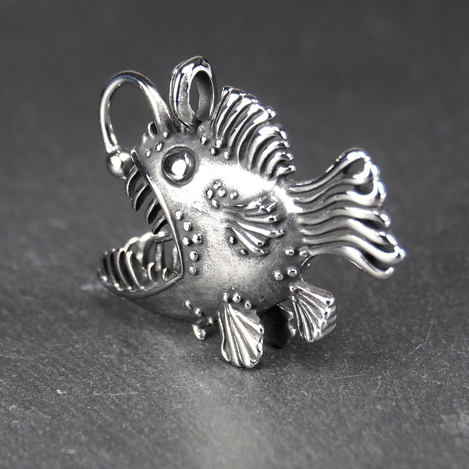 3D-Anhänger Anglerfisch, silberglänzend, mit geöffnetem Maul und dolchartigen Zähnen, geriffelten Flossen, punktiertem Körper und Öse.