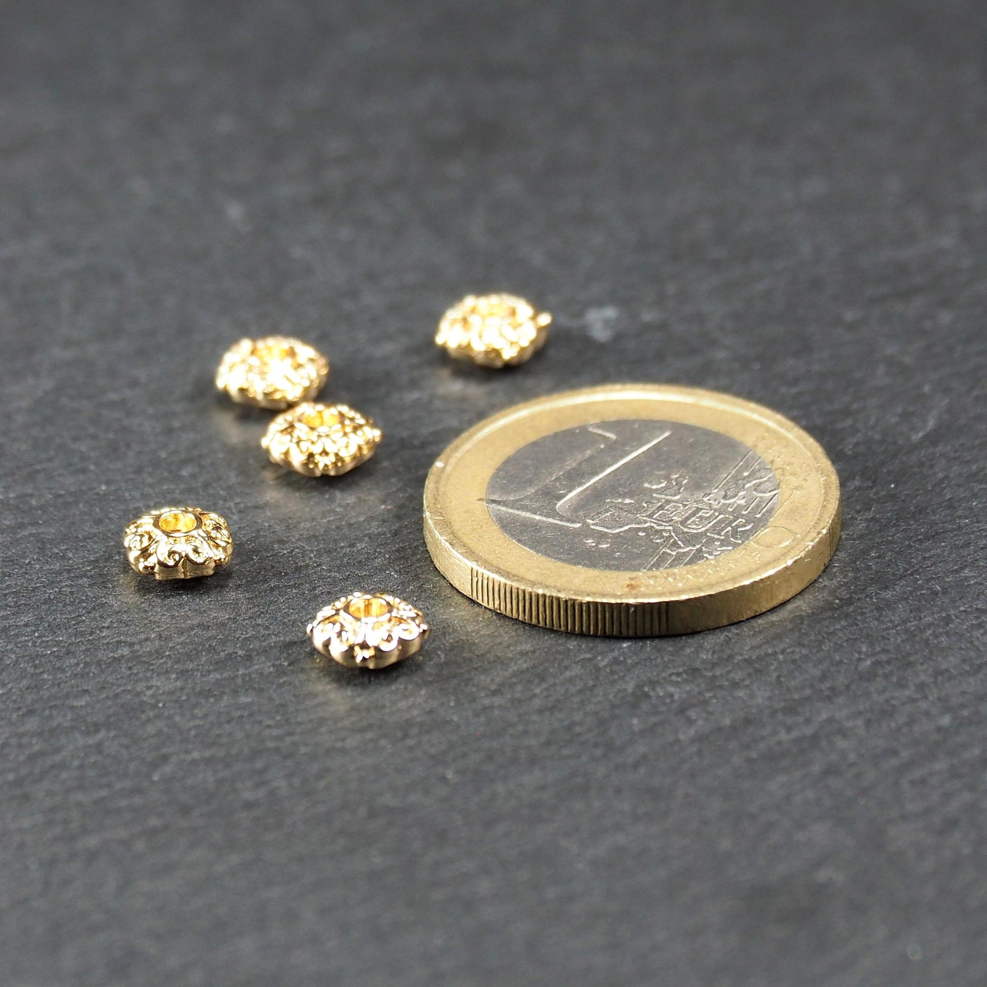 Runde goldfarbene Spacer Perlen mit glänzender Oberfläche, reliefartigem floralem Ornament und zentralem runden Durchbruch. Neben einer 1 Euro Münze zum Größenvergleich.
