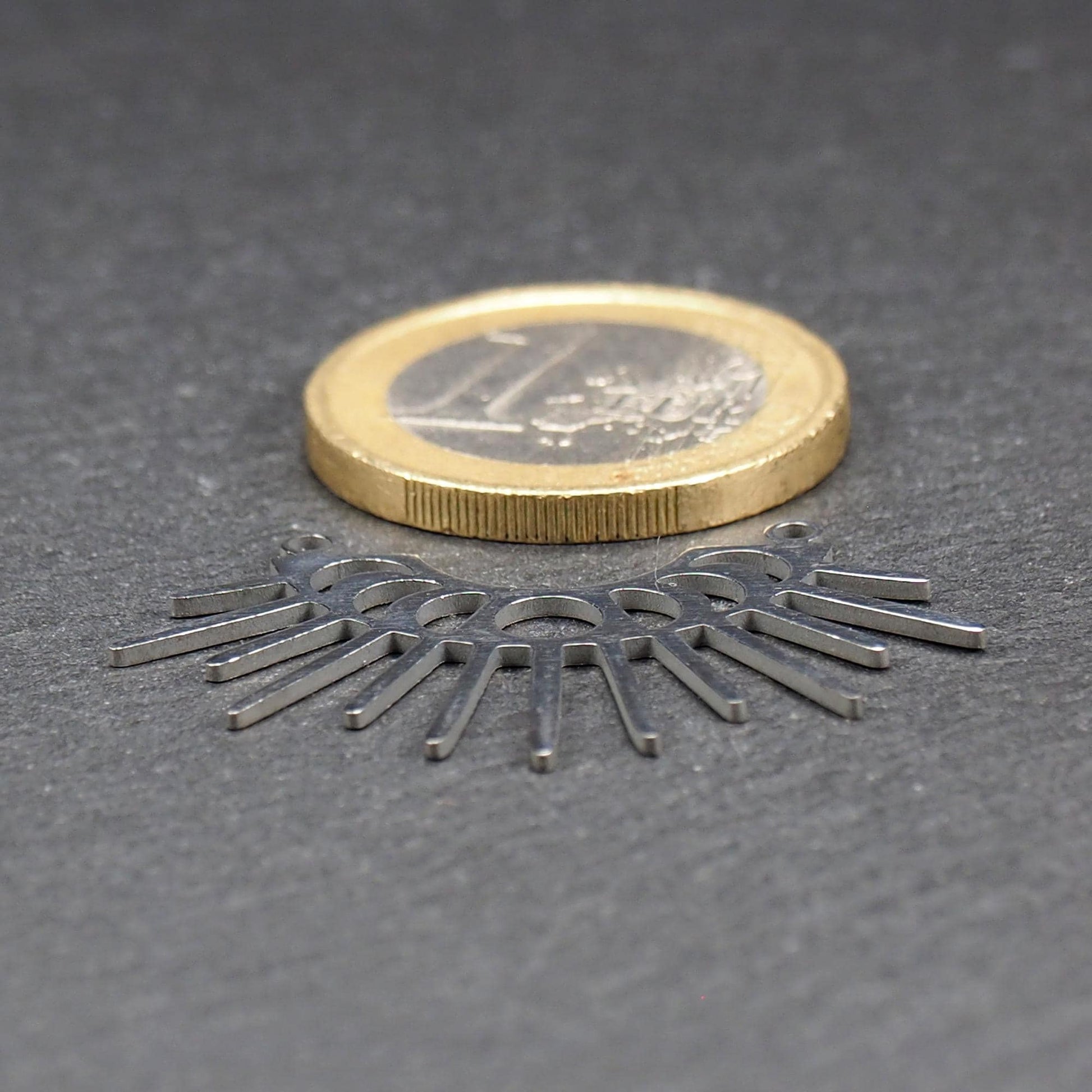 Versilberter Metallverbinder, halbkreisförmig und durchbrochen, mit rundem Mittelloch, mehreren schmalen radialen Strahlen, glänzender Oberfläche und zwei kleinen Ösen oben.