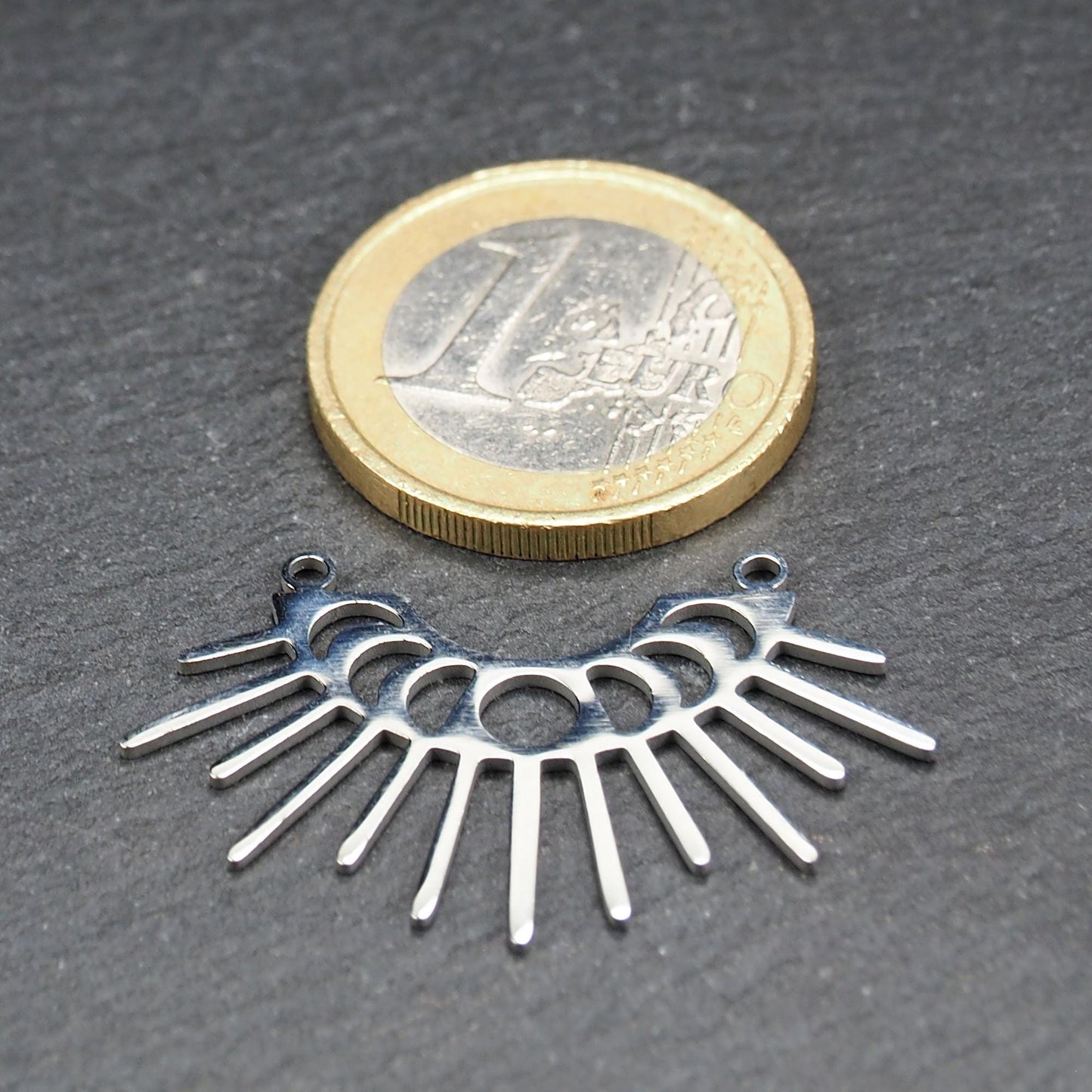Versilberter Metallverbinder, halbkreisförmig und durchbrochen, mit rundem Mittelloch, mehreren schmalen radialen Strahlen, glänzender Oberfläche und zwei kleinen Ösen oben.