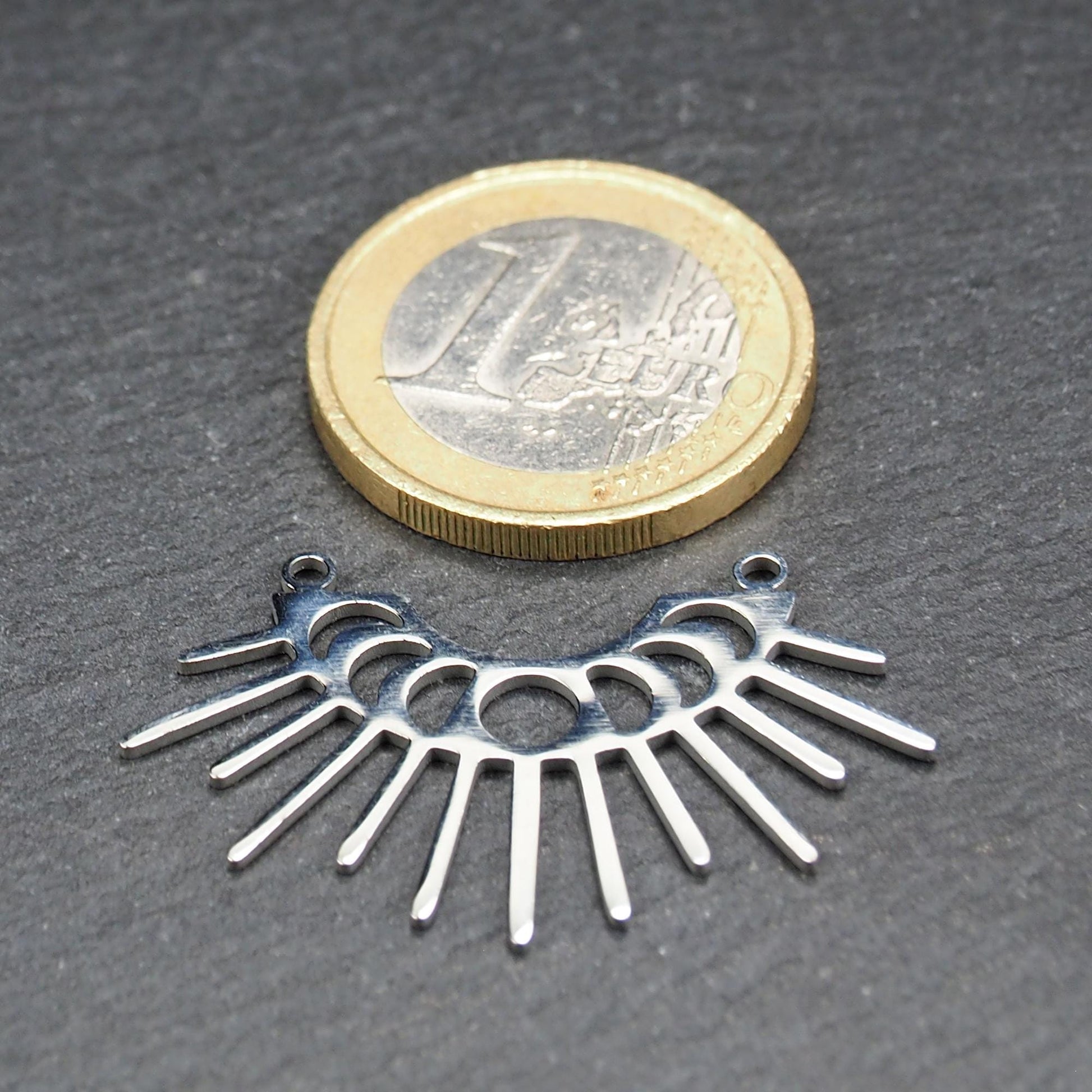 Versilberter Metallverbinder, halbkreisförmig und durchbrochen, mit rundem Mittelloch, mehreren schmalen radialen Strahlen, glänzender Oberfläche und zwei kleinen Ösen oben.