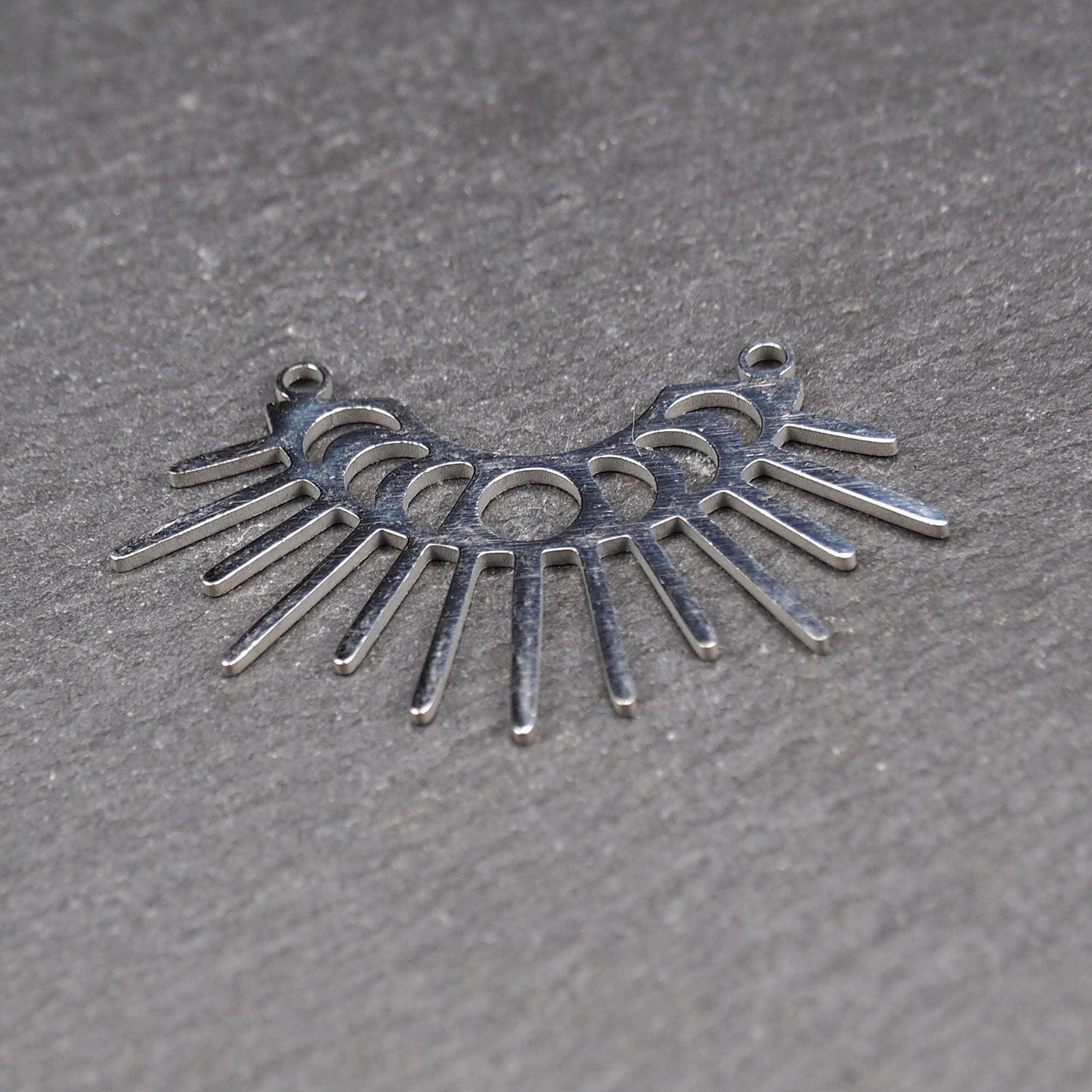 Versilberter Metallverbinder, halbkreisförmig und durchbrochen, mit rundem Mittelloch, mehreren schmalen radialen Strahlen, glänzender Oberfläche und zwei kleinen Ösen oben.