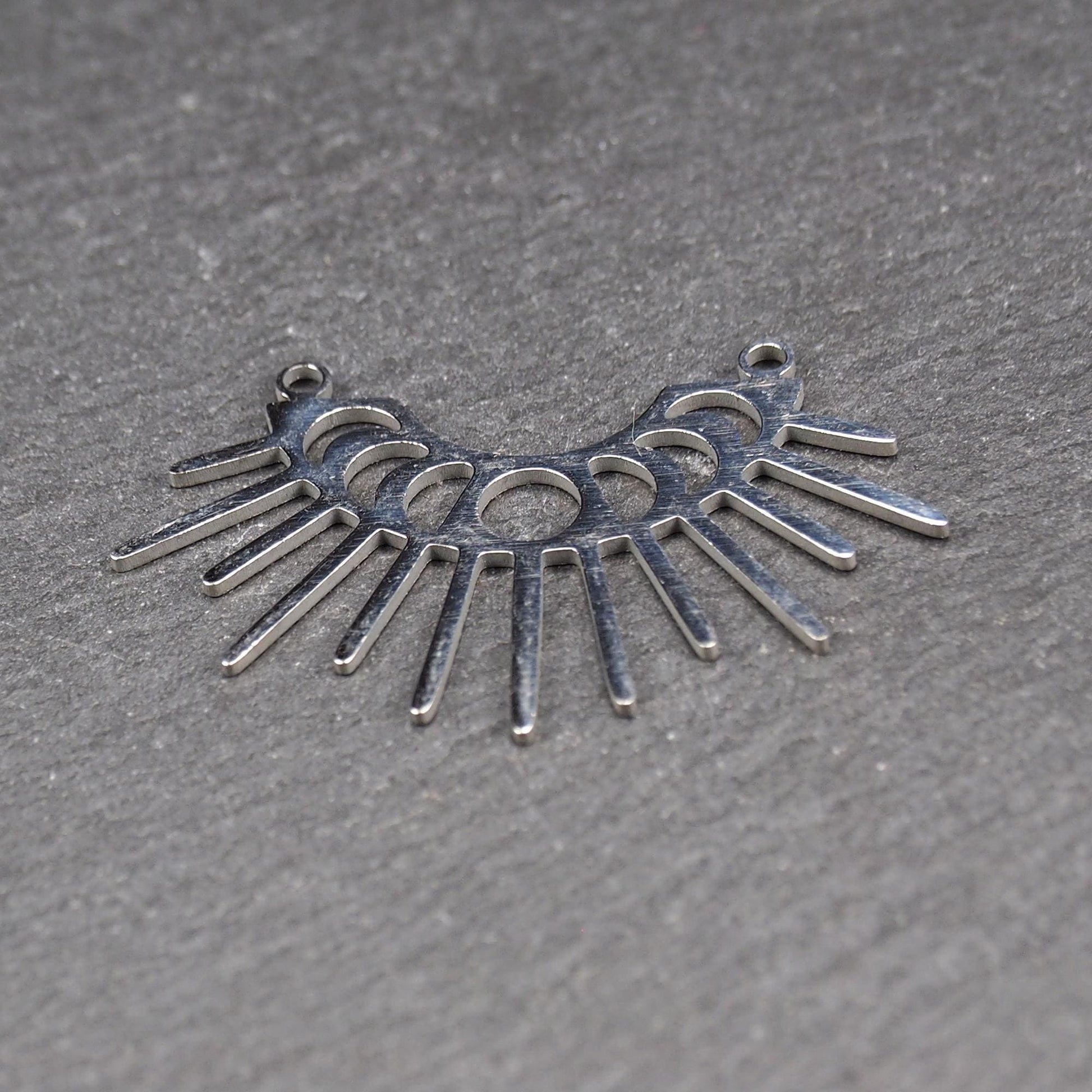 Versilberter Metallverbinder, halbkreisförmig und durchbrochen, mit rundem Mittelloch, mehreren schmalen radialen Strahlen, glänzender Oberfläche und zwei kleinen Ösen oben.