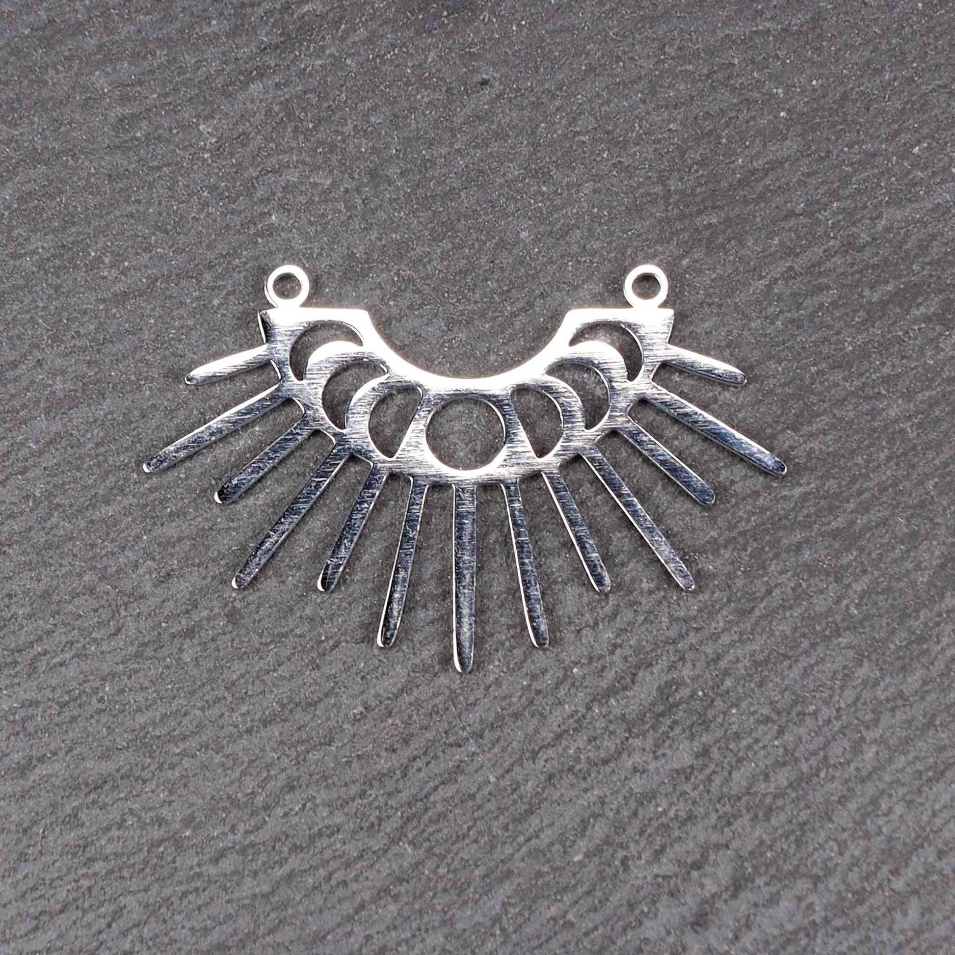 Versilberter Metallverbinder, halbkreisförmig und durchbrochen, mit rundem Mittelloch, mehreren schmalen radialen Strahlen, glänzender Oberfläche und zwei kleinen Ösen oben.