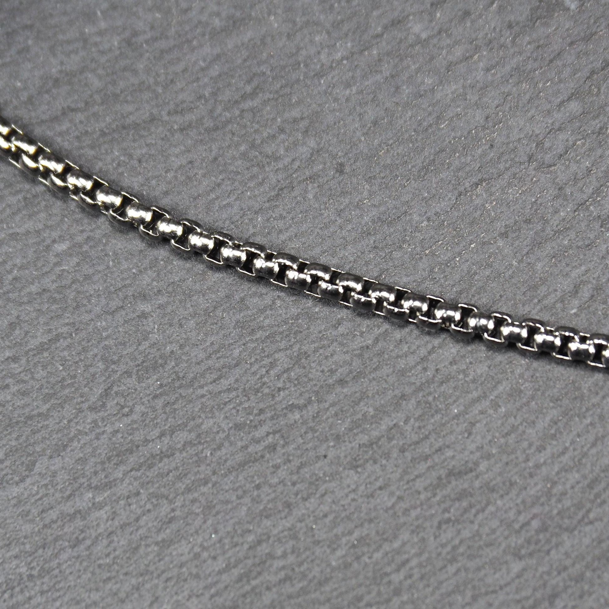 Quadratische Boxkette aus silberfarbenem, glänzendem Metall mit eng aneinandergereihten quadratischen Gliedern, diagonal gelegt.