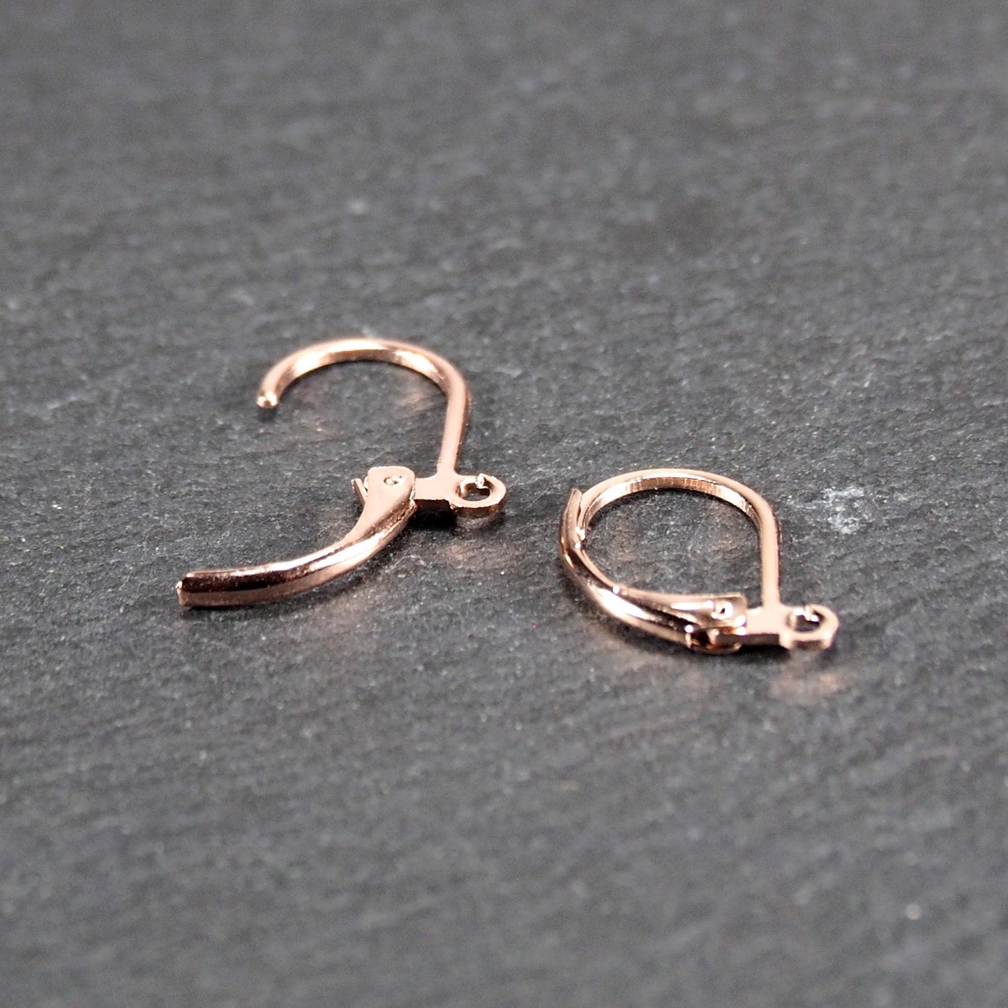 Zwei rosévergoldete Klappbrisuren in glänzender Metalloptik, ovale Leverback-Form mit sichtbarer kleinen Öse am unteren Ende, auf dunklem Hintergrund.