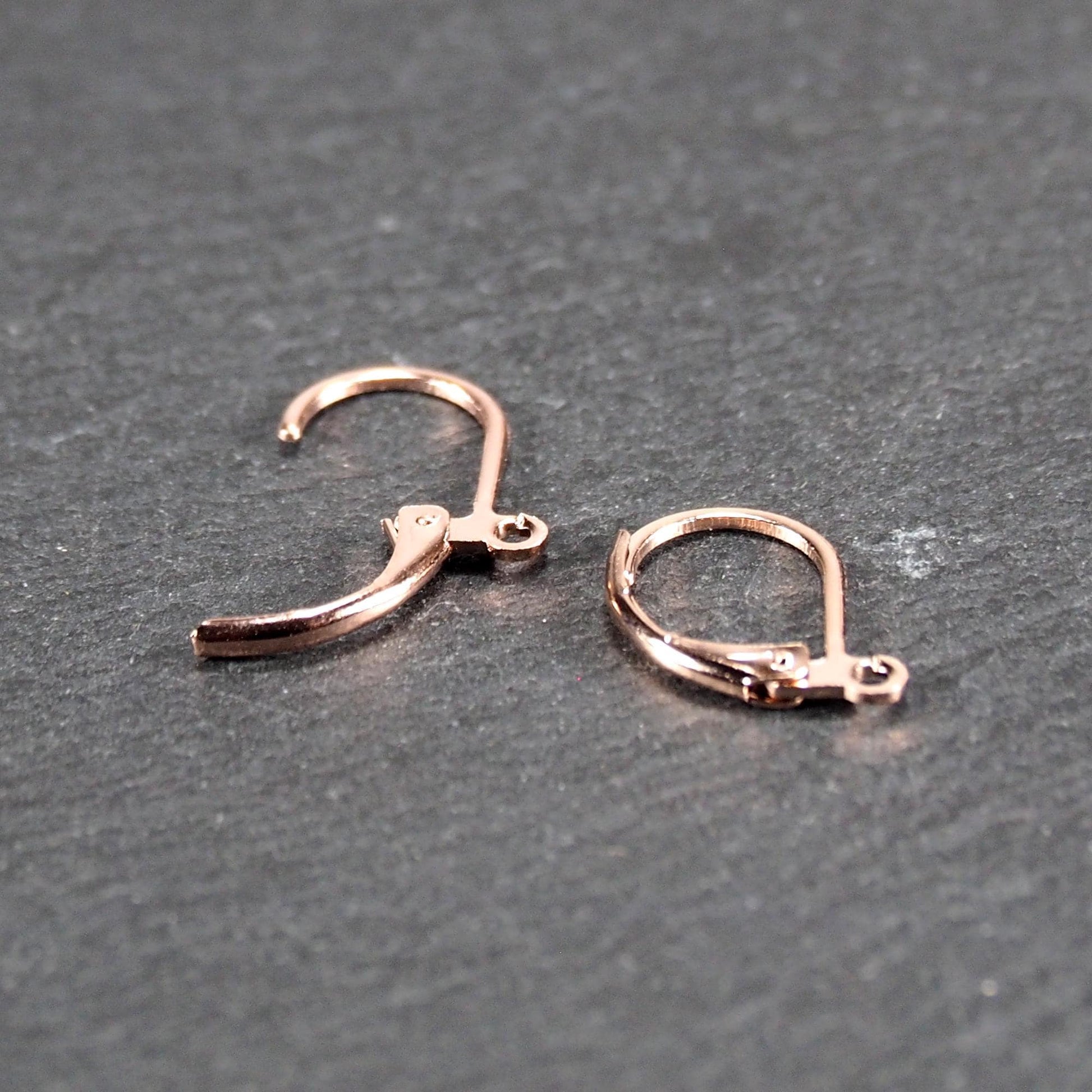 Zwei rosévergoldete Klappbrisuren in glänzender Metalloptik, ovale Leverback-Form mit sichtbarer kleinen Öse am unteren Ende, auf dunklem Hintergrund.