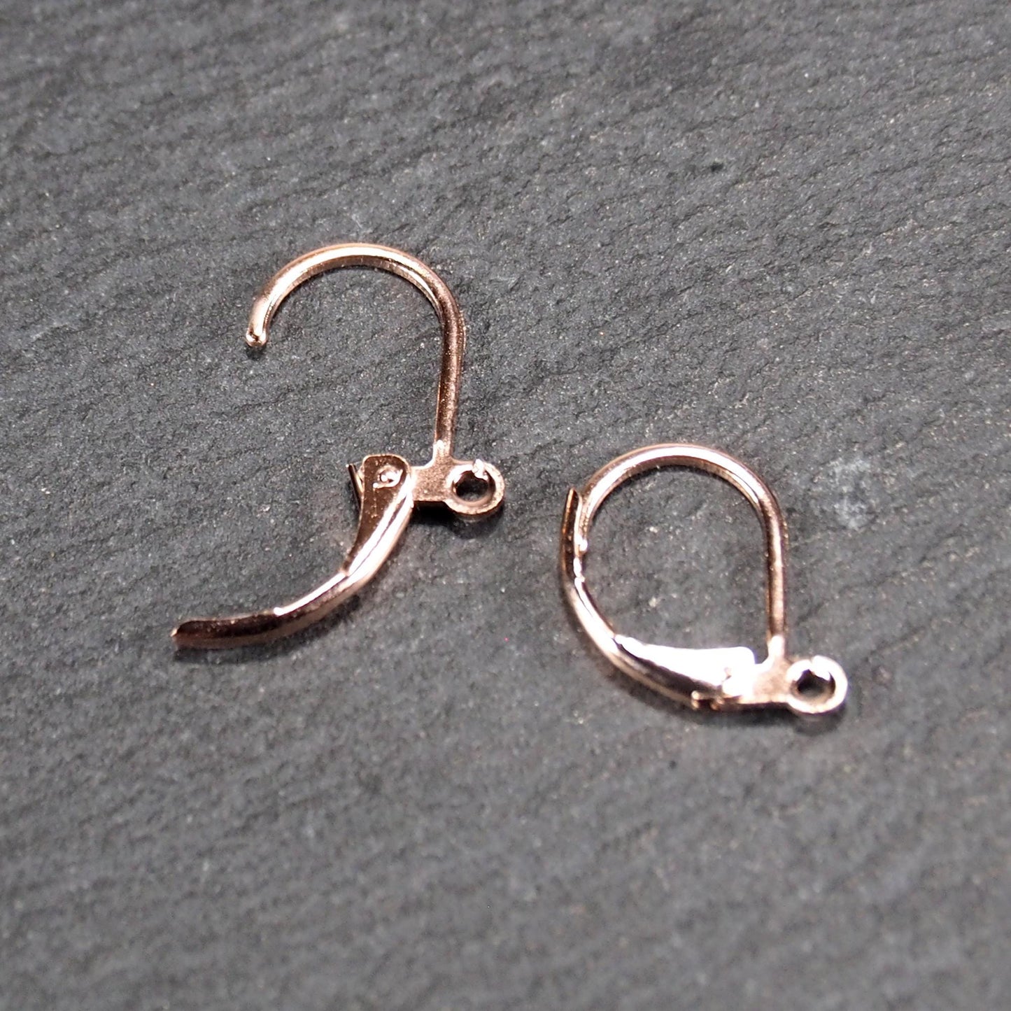 Zwei rosévergoldete Klappbrisuren in glänzender Metalloptik, ovale Leverback-Form mit sichtbarer kleinen Öse am unteren Ende, auf dunklem Hintergrund.