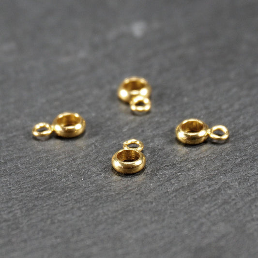 Vier Anhängerösen in goldfarbener, glänzender Ausführung, ringförmig mit kleiner seitlicher Öse auf dunklem Strukturhintergrund.