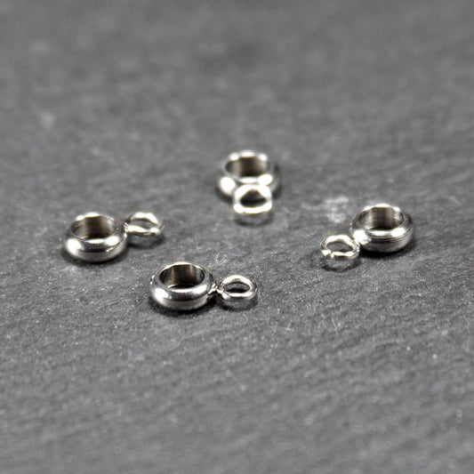 Vier runde, silberne, glänzende Anhängerösen in Donutform, jeweils mit einer kleinen seitlichen Öse, auf grauem Untergrund.