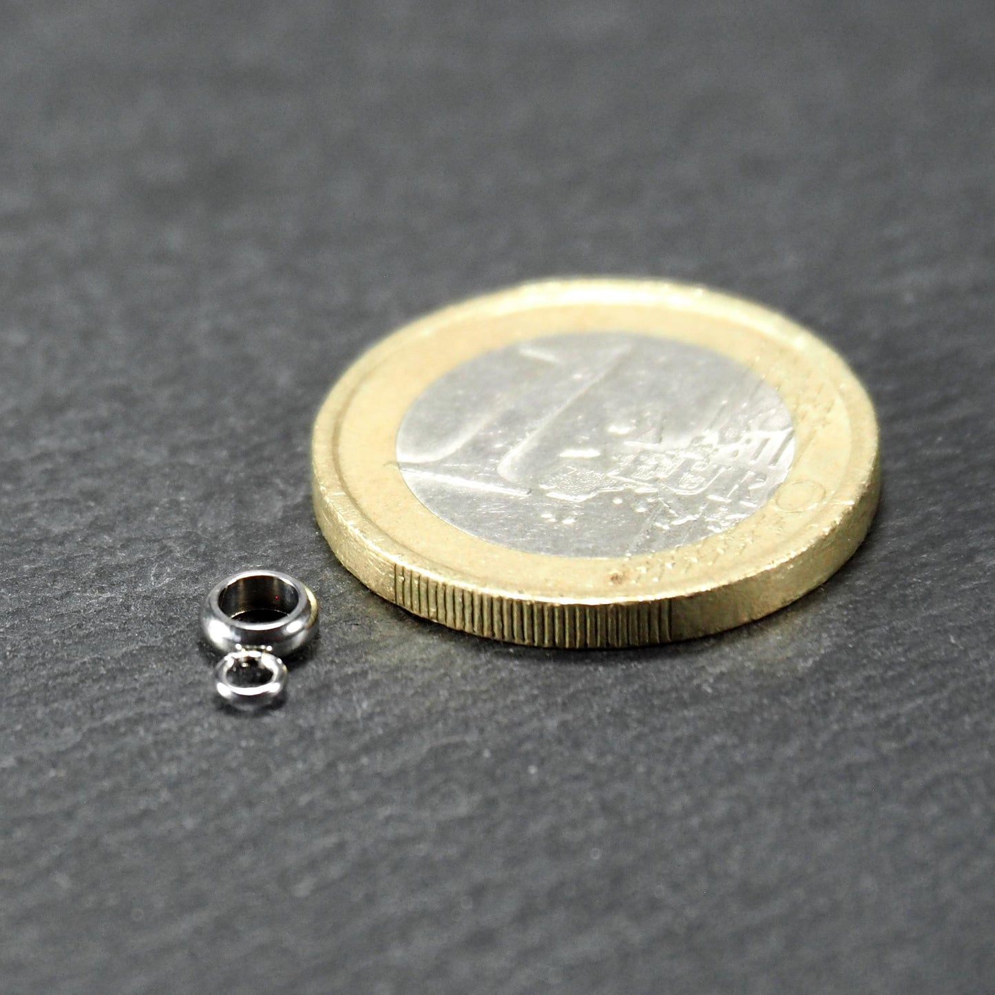 Kleine goldfarbene glänzende runde Anhängeröse in kompakter durchbrochener Ringform neben einer 1-Euro-Münze als Größenskalierung.