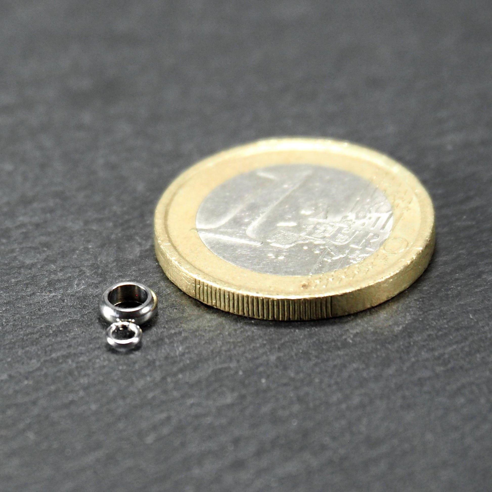 Kleine goldfarbene glänzende runde Anhängeröse in kompakter durchbrochener Ringform neben einer 1-Euro-Münze als Größenskalierung.
