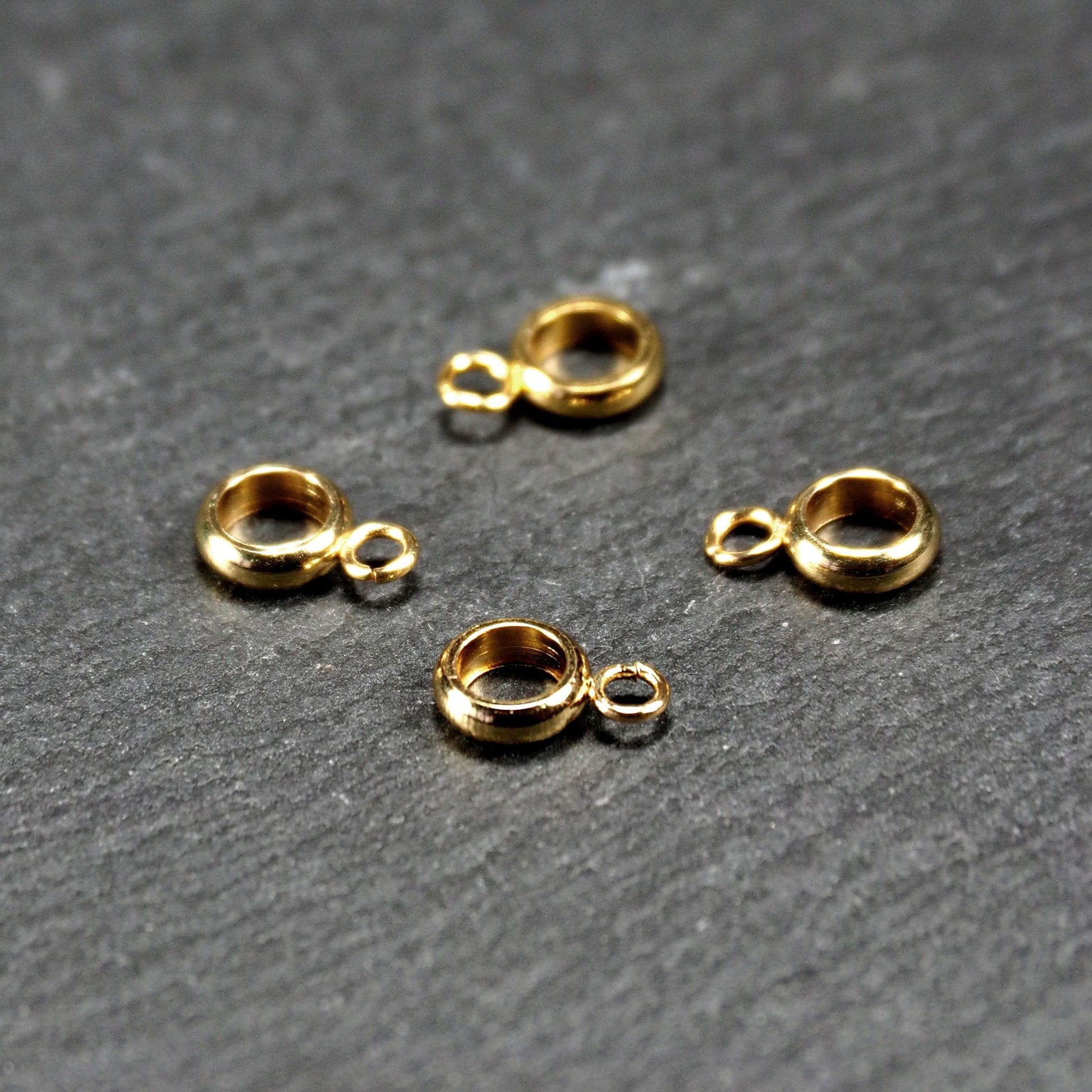 Vier vergoldete ringförmige Anhängerösen mit glänzender goldfarbener Oberfläche, hohlem Mittelring und jeweils kleinem seitlichem Ösenring.