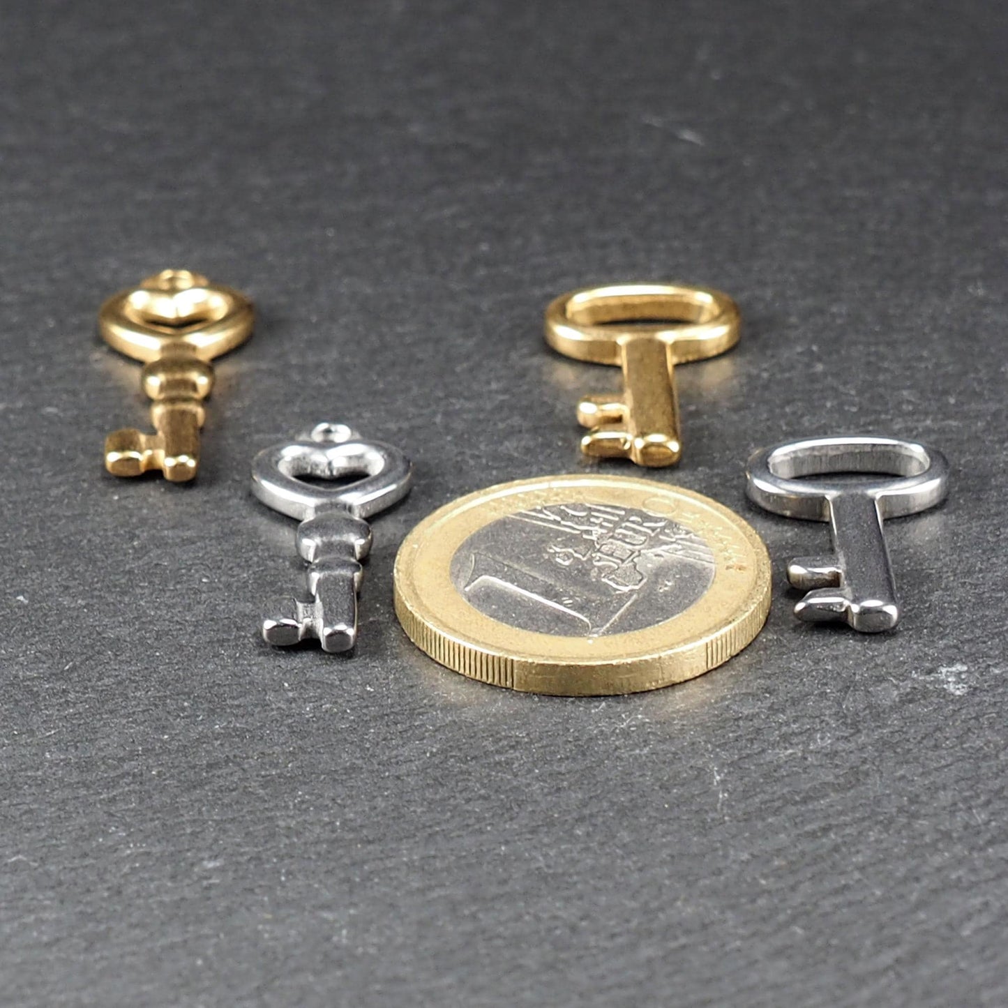Vier kleine, glänzende schlüsselförmige Anhänger in Silber und Gold mit herzförmigen Ösen, neben einer 1-Euro-Münze.