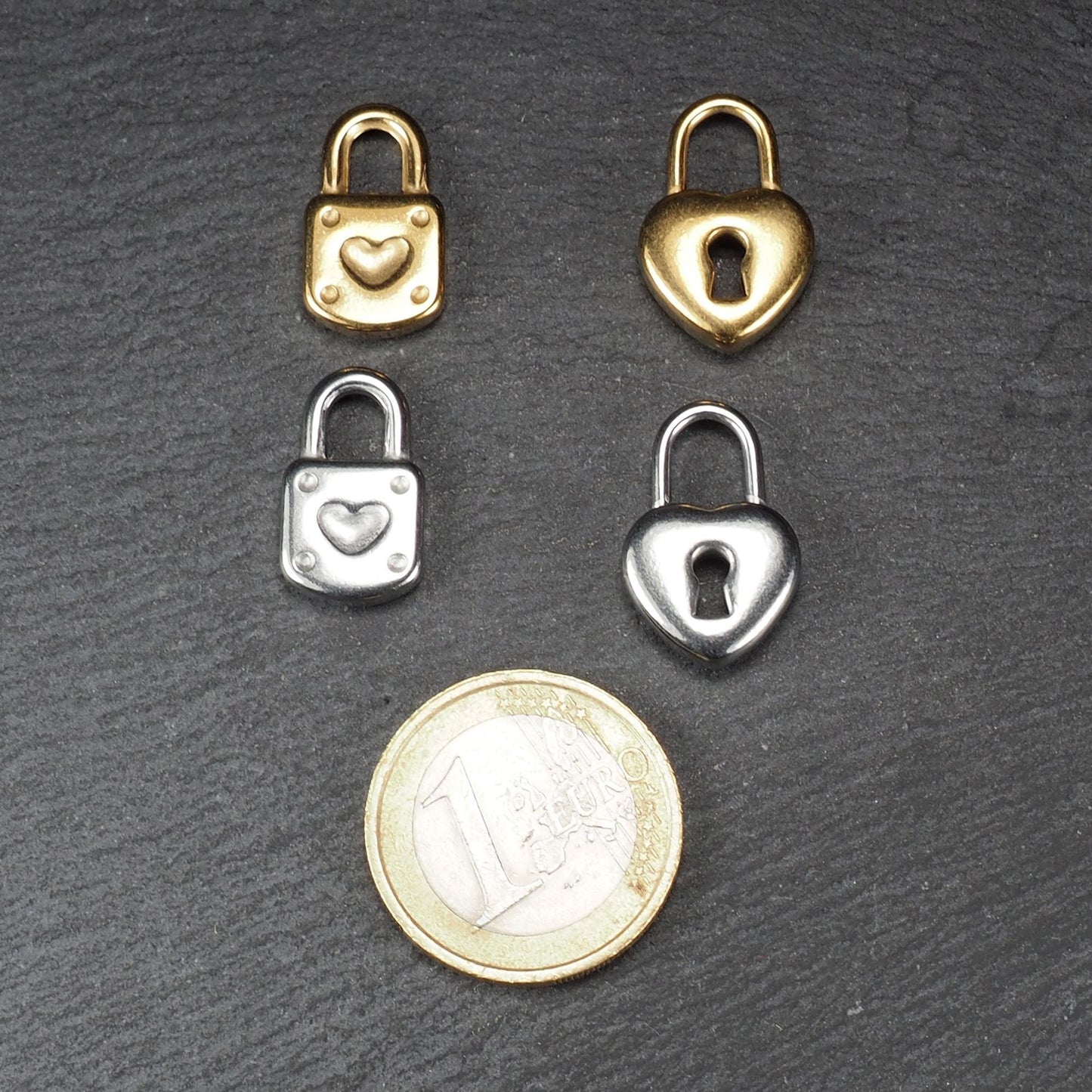 Vier kleine Anhänger in Schloss- und Herzform, glatt glänzend, zwei vergoldet, zwei silberfarben, mit sichtbarer Öse, neben einer 1‑Euro‑Münze.