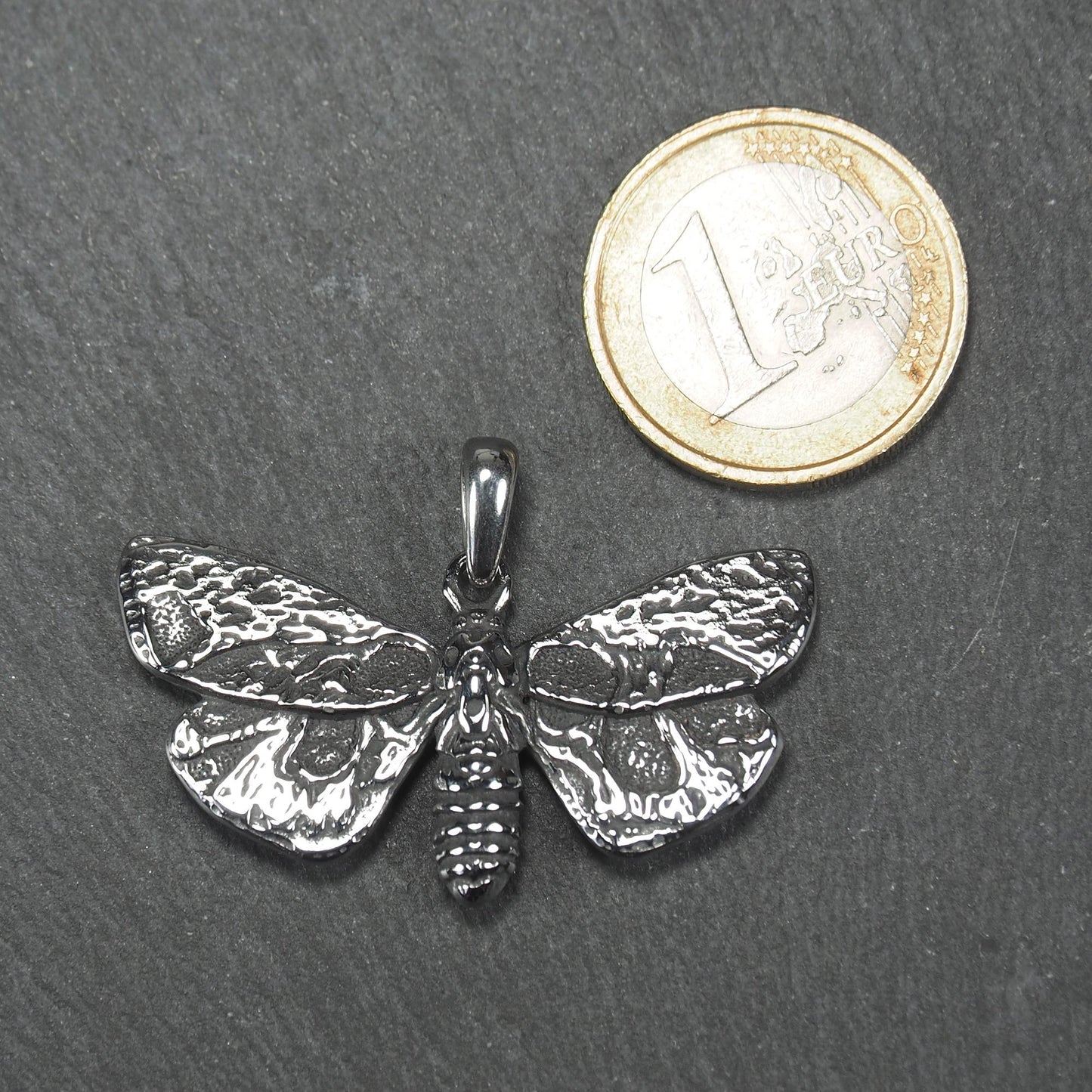 Anhänger in Mottenform, silberfarbene glänzende Metalloberfläche mit reliefartigen, strukturierten Flügeln, segmentiertem Körper und integrierter Öse. Mit 1 Euro Münze zum Größenvergleich.