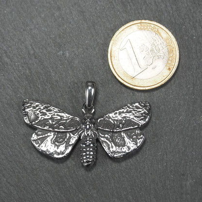 Anhänger in Mottenform, silberfarbene glänzende Metalloberfläche mit reliefartigen, strukturierten Flügeln, segmentiertem Körper und integrierter Öse. Mit 1 Euro Münze zum Größenvergleich.