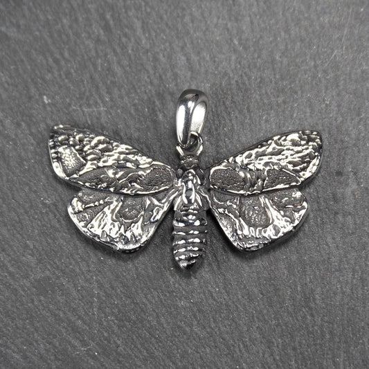 Anhänger in Mottenform, silberfarbene glänzende Metalloberfläche mit reliefartigen, strukturierten Flügeln, segmentiertem Körper und integrierter Öse.
