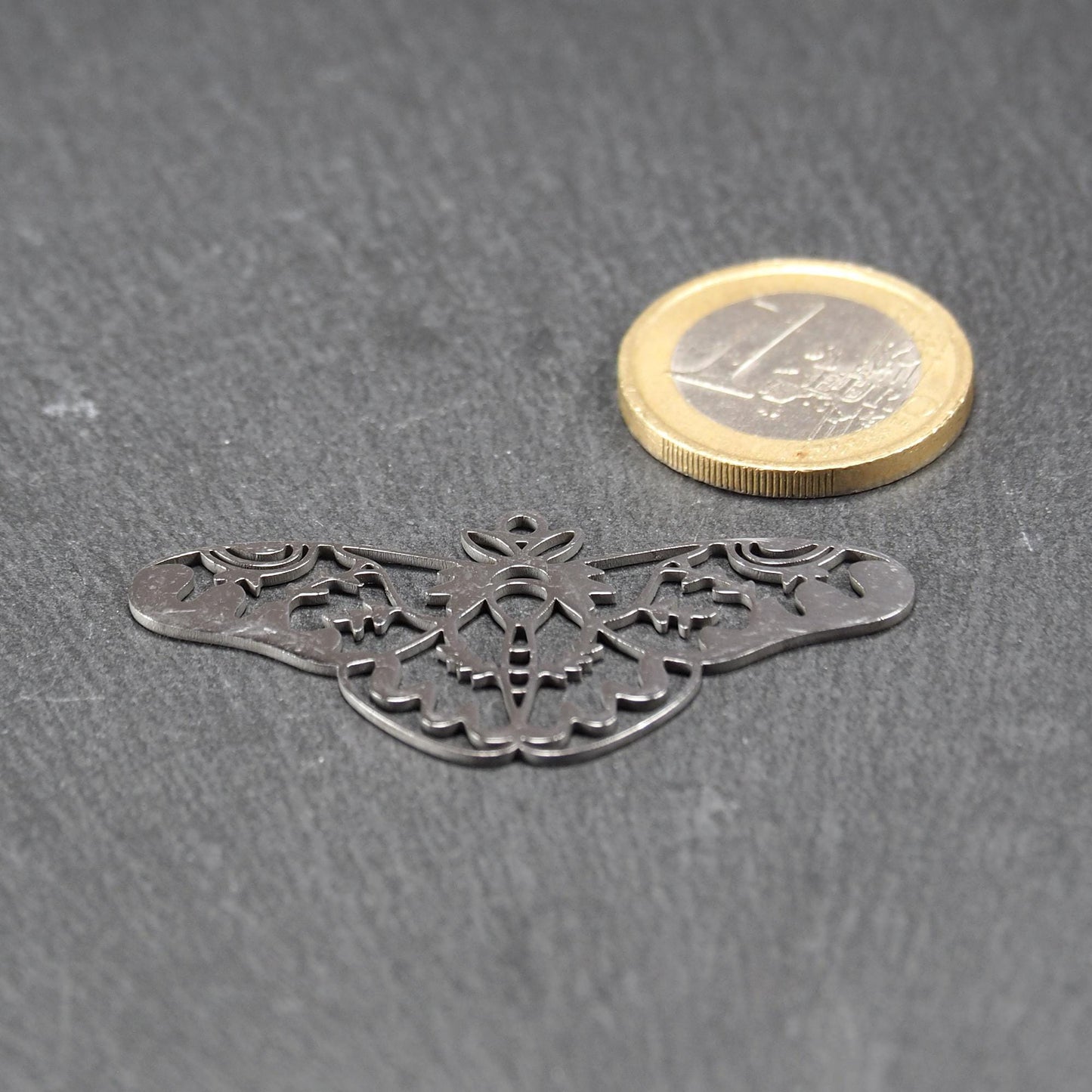 Anhänger / Verbinder in Mottenform aus poliertem silberfarbenem Edelstahl, filigranes durchbrochenes Ornament-Flügeldesign mit integrierter Öse oben. Neben einer 1 Euro Münze zum Größenvergleich.