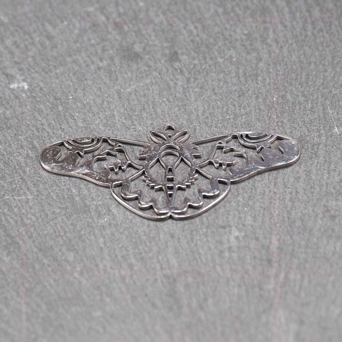 Anhänger / Verbinder in Mottenform aus poliertem silberfarbenem Edelstahl, filigranes durchbrochenes Ornament-Flügeldesign mit integrierter Öse oben.
