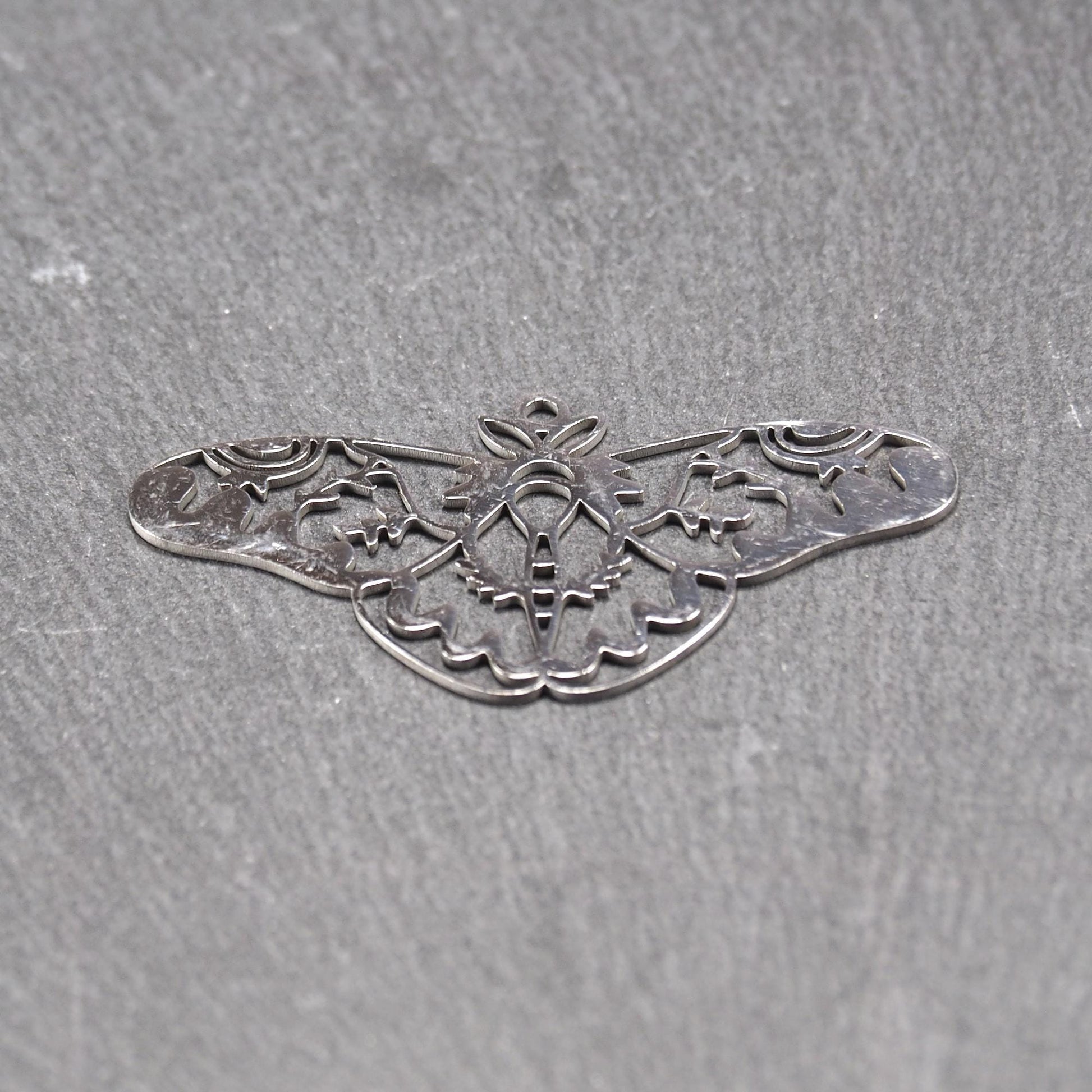 Anhänger / Verbinder in Mottenform aus poliertem silberfarbenem Edelstahl, filigranes durchbrochenes Ornament-Flügeldesign mit integrierter Öse oben.