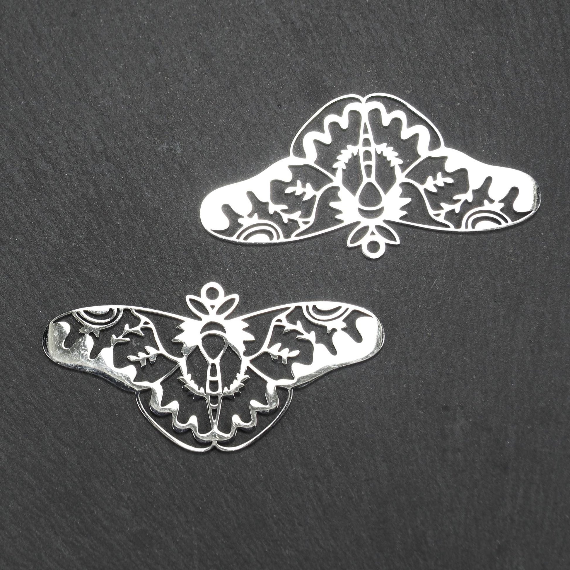 Anhänger / Verbinder in Mottenform aus poliertem silberfarbenem Edelstahl, filigranes durchbrochenes Ornament-Flügeldesign mit integrierter Öse oben.