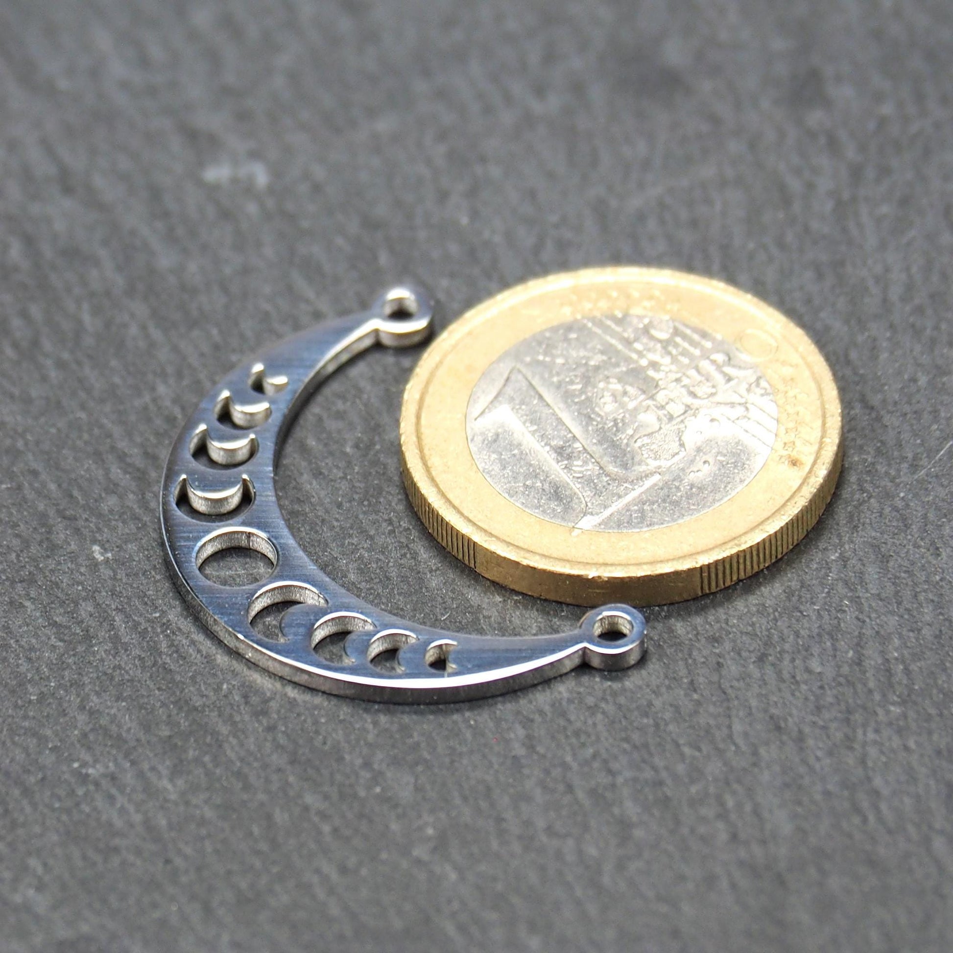 Sichelförmiger silberner Metallverbinder mit ausgeschnittenem Mondphasen-Motiv und je einer Rundöse an den Enden.