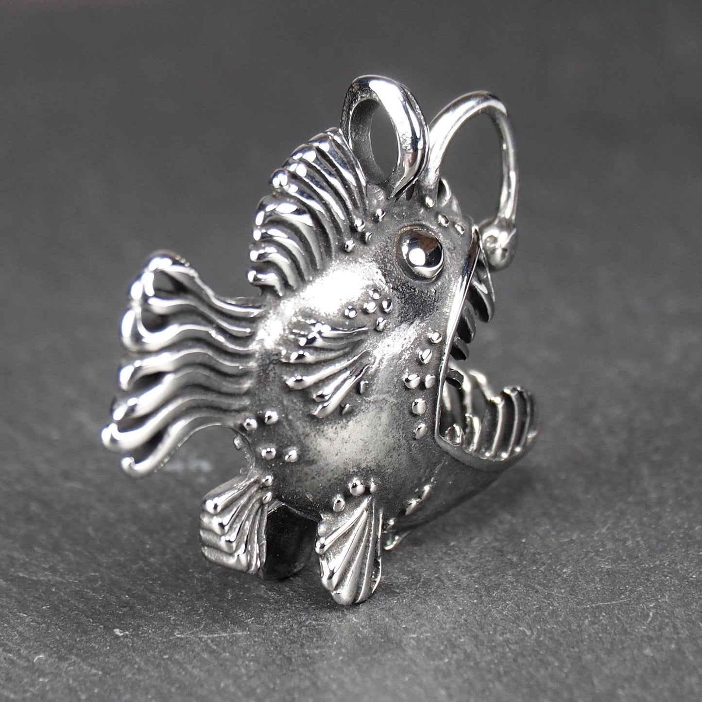 3D-Anhänger Anglerfisch, silberglänzend, mit geöffnetem Maul und dolchartigen Zähnen, geriffelten Flossen, punktiertem Körper und Öse.