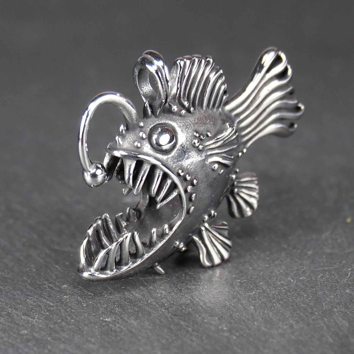 3D-Anhänger Anglerfisch, silberglänzend, mit geöffnetem Maul und dolchartigen Zähnen, geriffelten Flossen, punktiertem Körper und Öse.