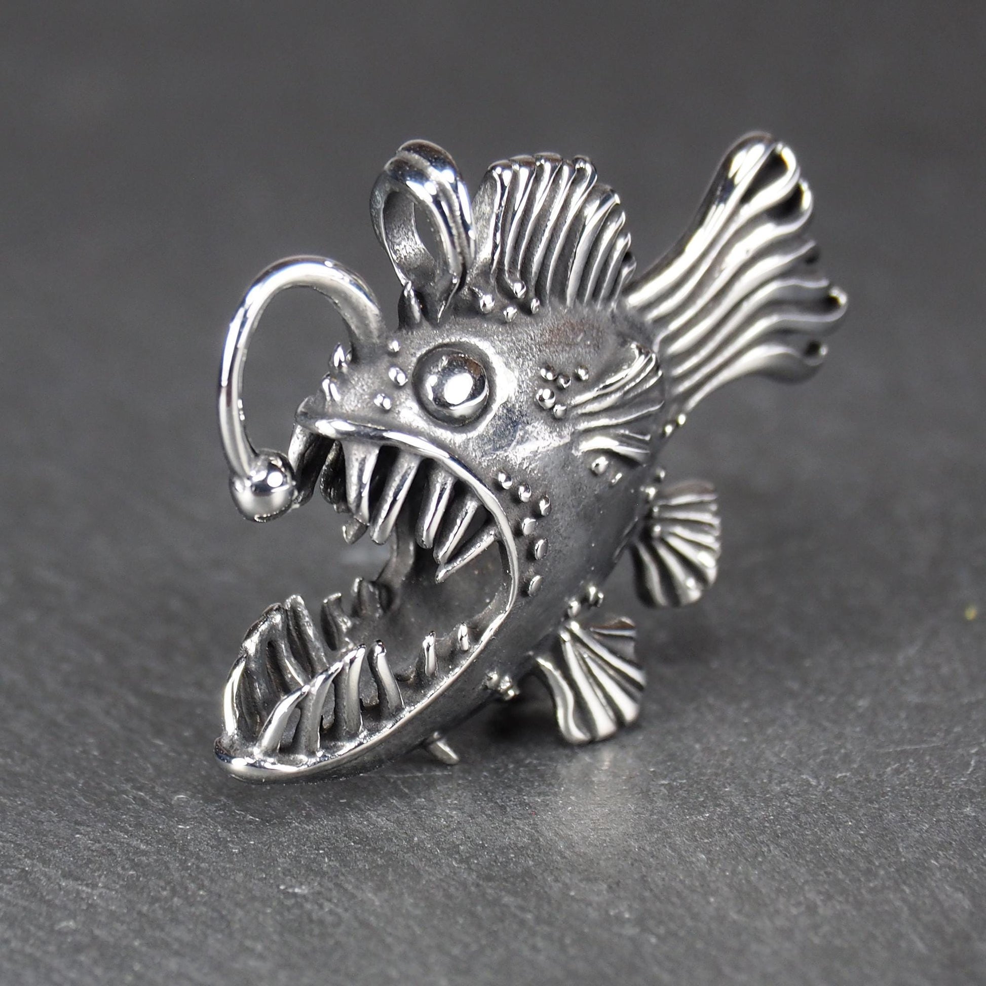 3D-Anhänger Anglerfisch, silberglänzend, mit geöffnetem Maul und dolchartigen Zähnen, geriffelten Flossen, punktiertem Körper und Öse.