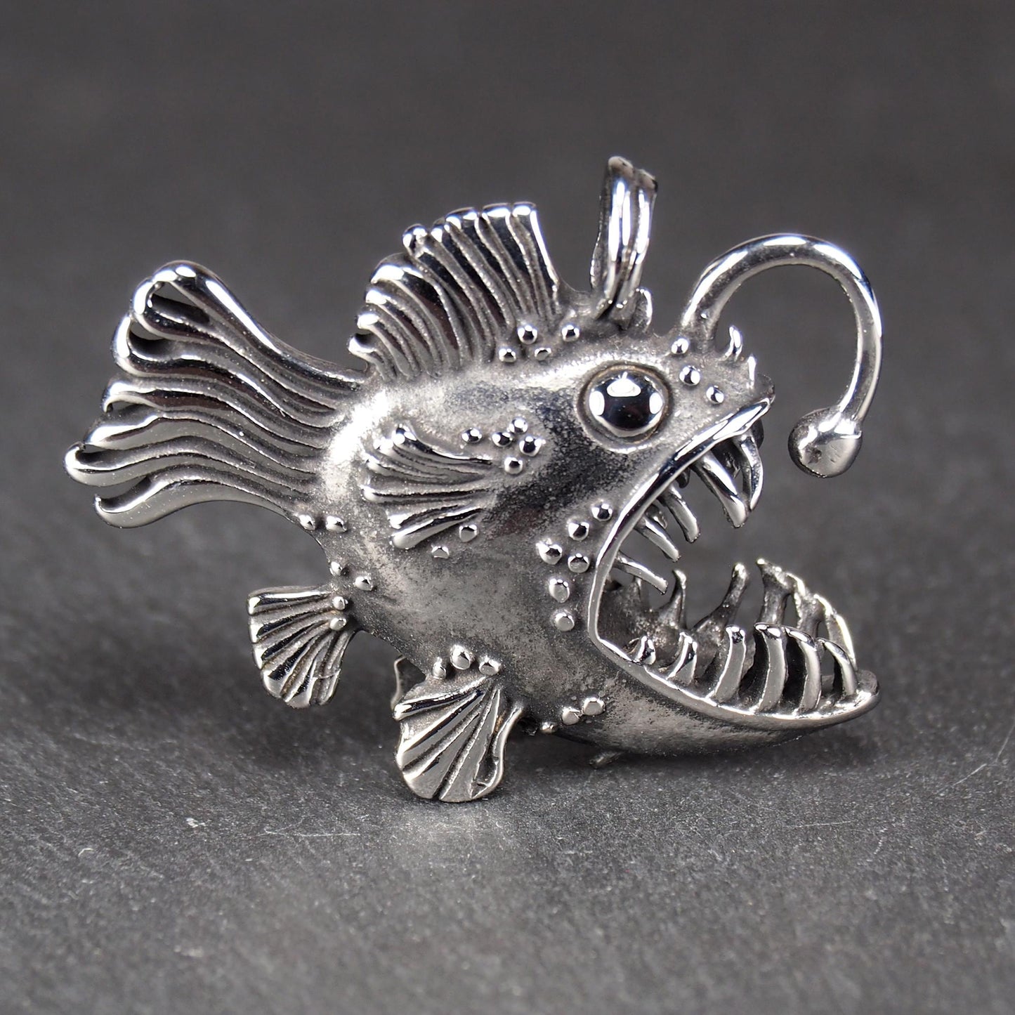 3D-Anhänger Anglerfisch, silberglänzend, mit geöffnetem Maul und dolchartigen Zähnen, geriffelten Flossen, punktiertem Körper und Öse.