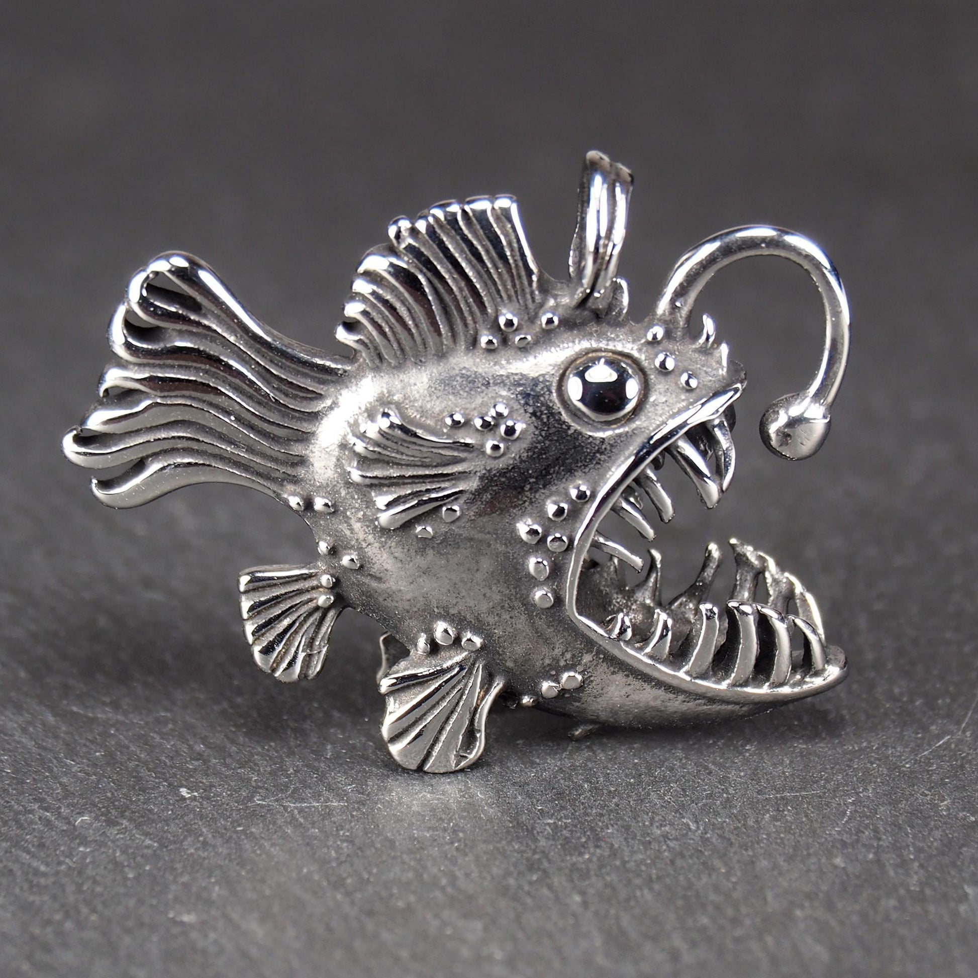3D-Anhänger Anglerfisch, silberglänzend, mit geöffnetem Maul und dolchartigen Zähnen, geriffelten Flossen, punktiertem Körper und Öse.