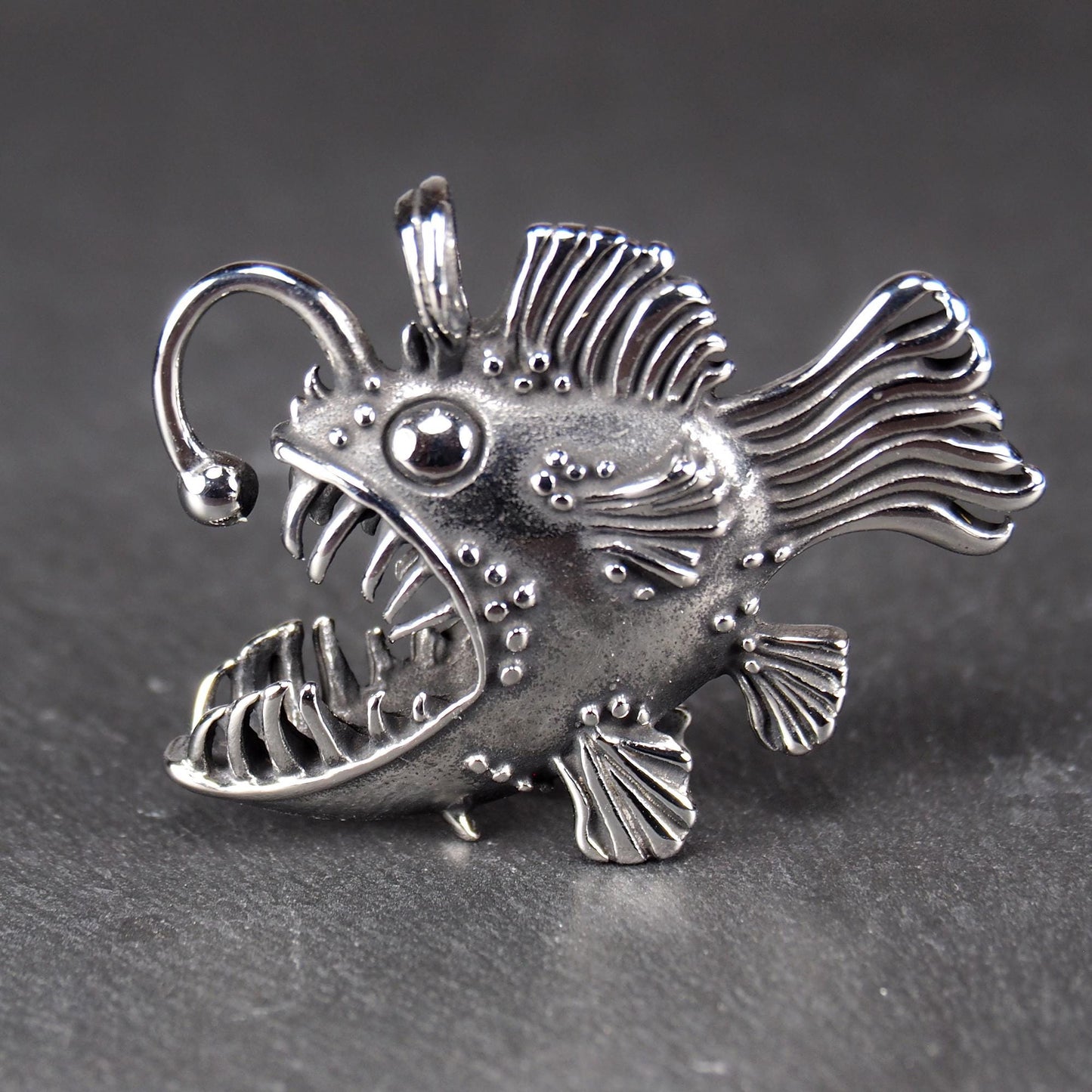 3D-Anhänger Anglerfisch, silberglänzend, mit geöffnetem Maul und dolchartigen Zähnen, geriffelten Flossen, punktiertem Körper und Öse.
