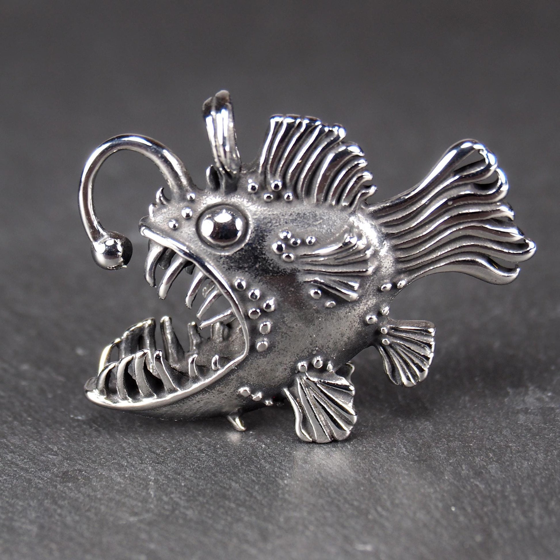 3D-Anhänger Anglerfisch, silberglänzend, mit geöffnetem Maul und dolchartigen Zähnen, geriffelten Flossen, punktiertem Körper und Öse.