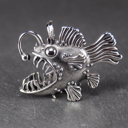 3D-Anhänger Anglerfisch, silberglänzend, mit geöffnetem Maul und dolchartigen Zähnen, geriffelten Flossen, punktiertem Körper und Öse.