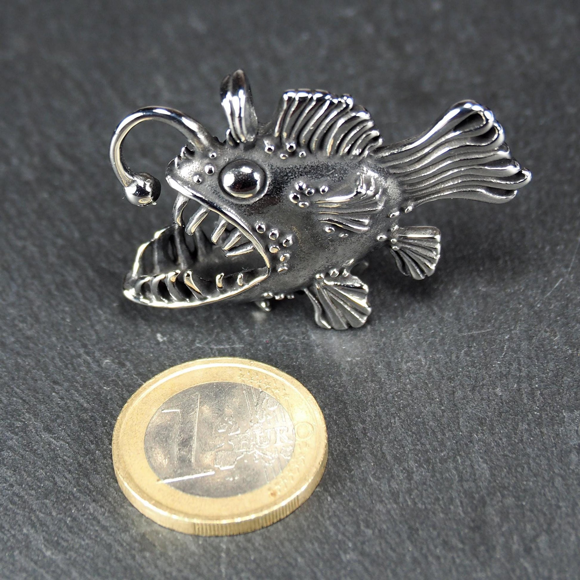 3D-Anhänger Anglerfisch, silberglänzend, mit geöffnetem Maul und dolchartigen Zähnen, geriffelten Flossen, punktiertem Körper und Öse. Daneben eine 1 Euro Münze zum Größenvergleich.