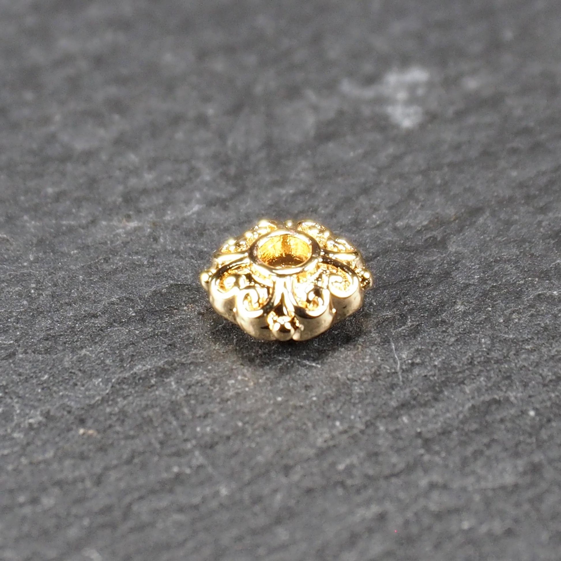Runde goldfarbene Spacer Perle mit glänzender Oberfläche, reliefartigem floralem Ornament und zentralem runden Durchbruch.
