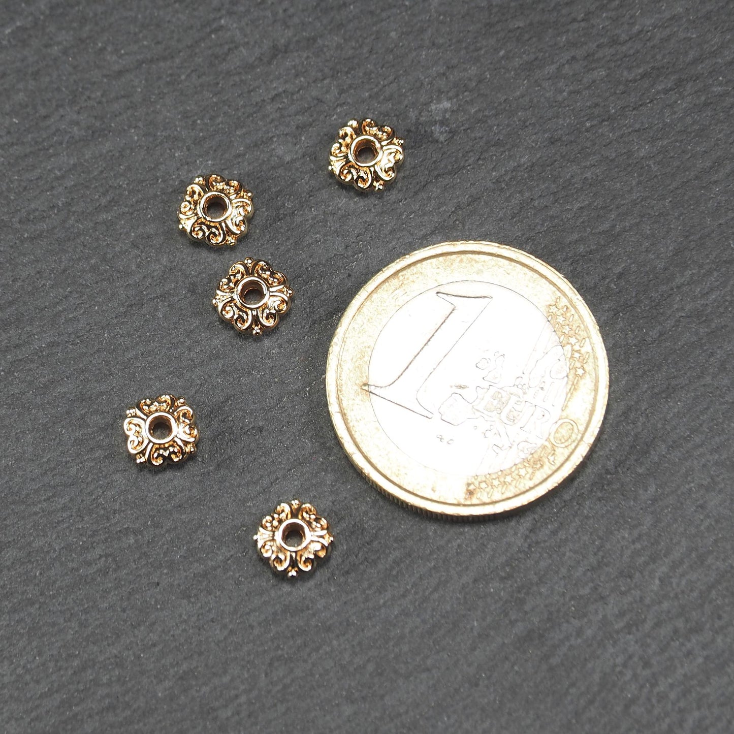 Runde goldfarbene Spacer Perlen mit glänzender Oberfläche, reliefartigem floralem Ornament und zentralem runden Durchbruch. Neben einer 1 Euro Münze zum Größenvergleich.