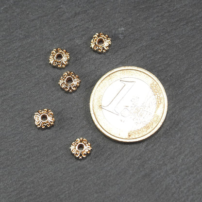 Runde goldfarbene Spacer Perlen mit glänzender Oberfläche, reliefartigem floralem Ornament und zentralem runden Durchbruch. Neben einer 1 Euro Münze zum Größenvergleich.