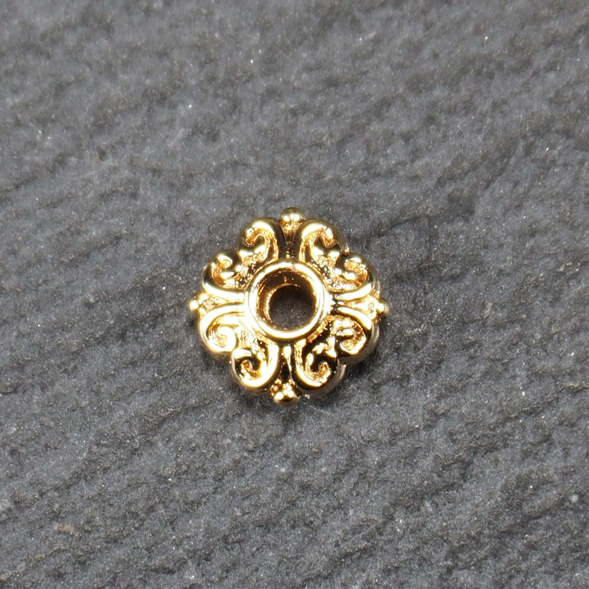 Runde goldfarbene Spacer Perle mit glänzender Oberfläche, reliefartigem floralem Ornament und zentralem runden Durchbruch.