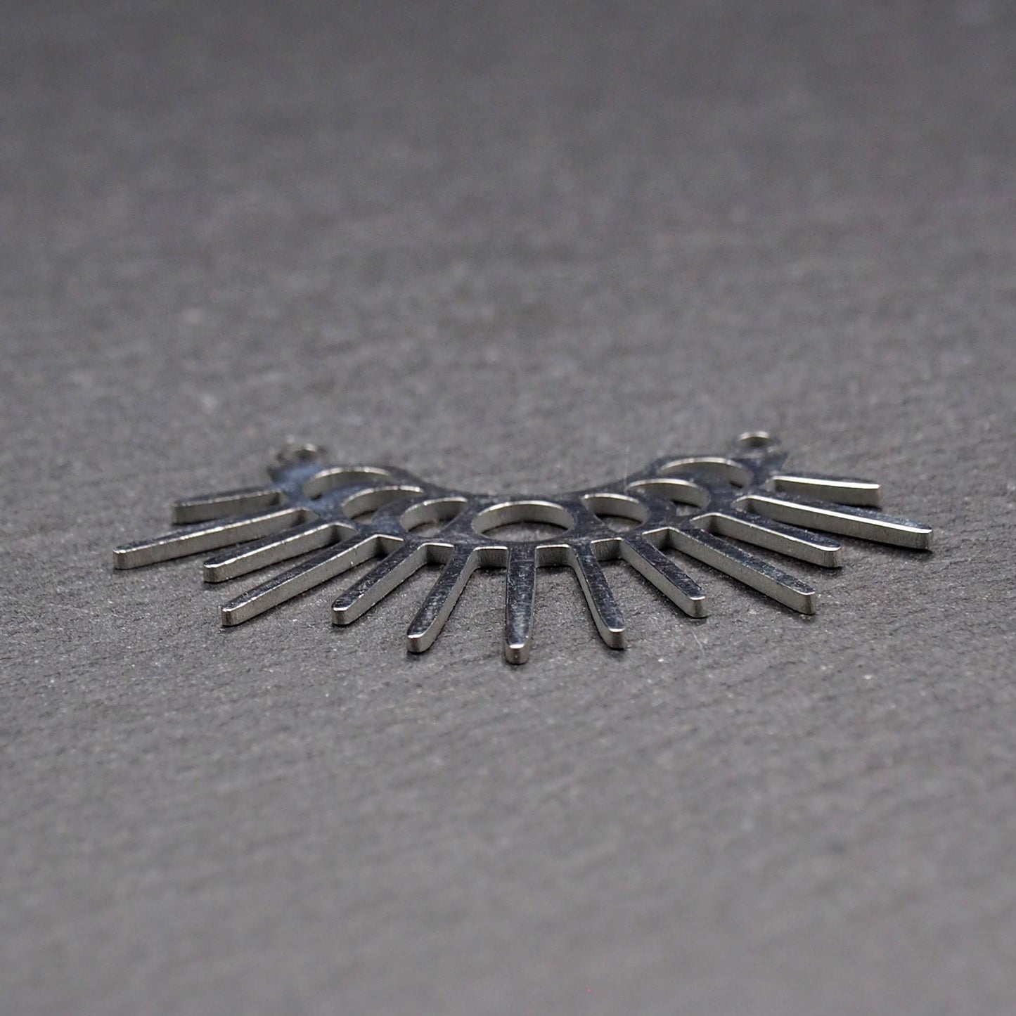 Versilberter Metallverbinder, halbkreisförmig und durchbrochen, mit rundem Mittelloch, mehreren schmalen radialen Strahlen, glänzender Oberfläche und zwei kleinen Ösen oben.