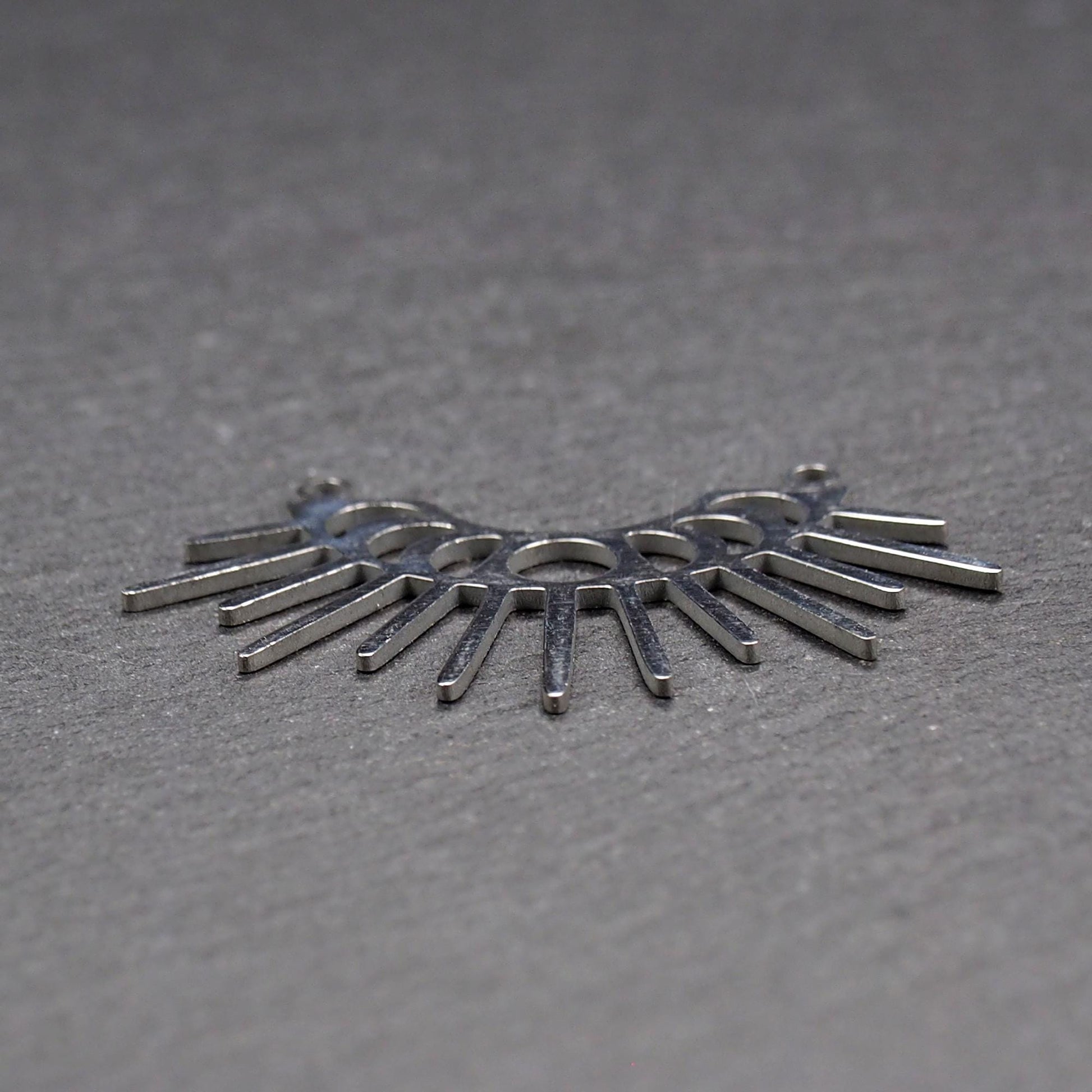 Versilberter Metallverbinder, halbkreisförmig und durchbrochen, mit rundem Mittelloch, mehreren schmalen radialen Strahlen, glänzender Oberfläche und zwei kleinen Ösen oben.