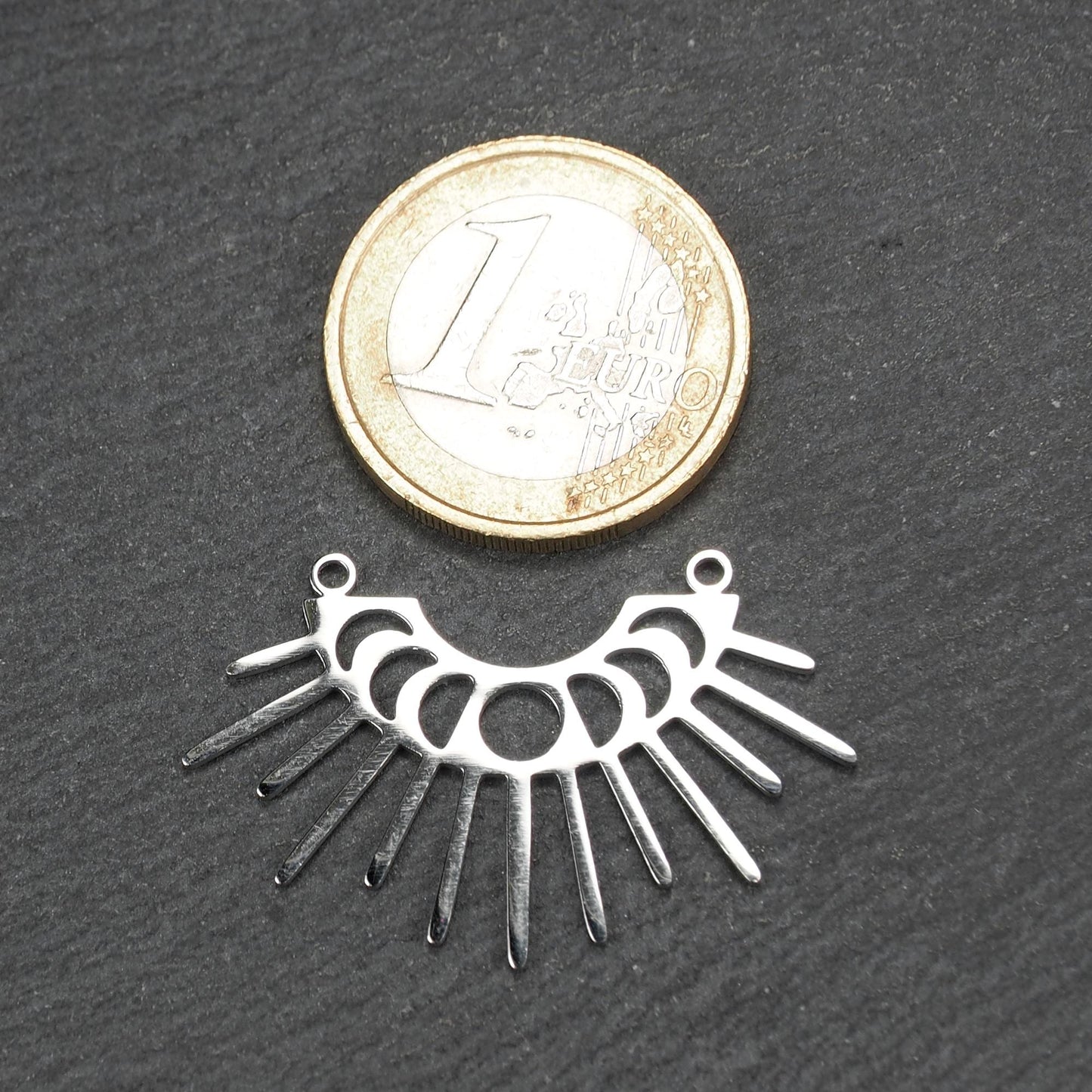 Versilberter Metallverbinder, halbkreisförmig und durchbrochen, mit rundem Mittelloch, mehreren schmalen radialen Strahlen, glänzender Oberfläche und zwei kleinen Ösen oben.
