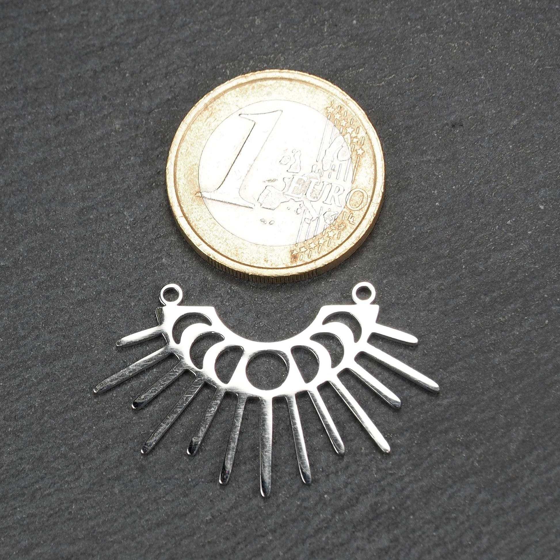 Versilberter Metallverbinder, halbkreisförmig und durchbrochen, mit rundem Mittelloch, mehreren schmalen radialen Strahlen, glänzender Oberfläche und zwei kleinen Ösen oben.